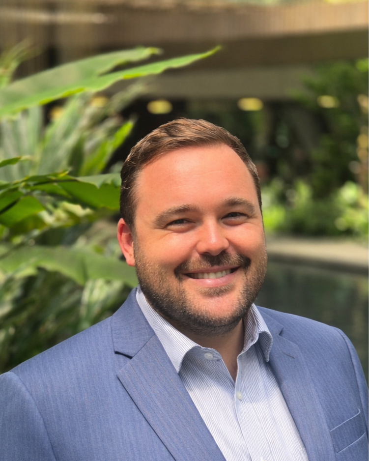 A man smiling in a blue suit and light-colored shirt outdoors with green plants and a body of water in the background.