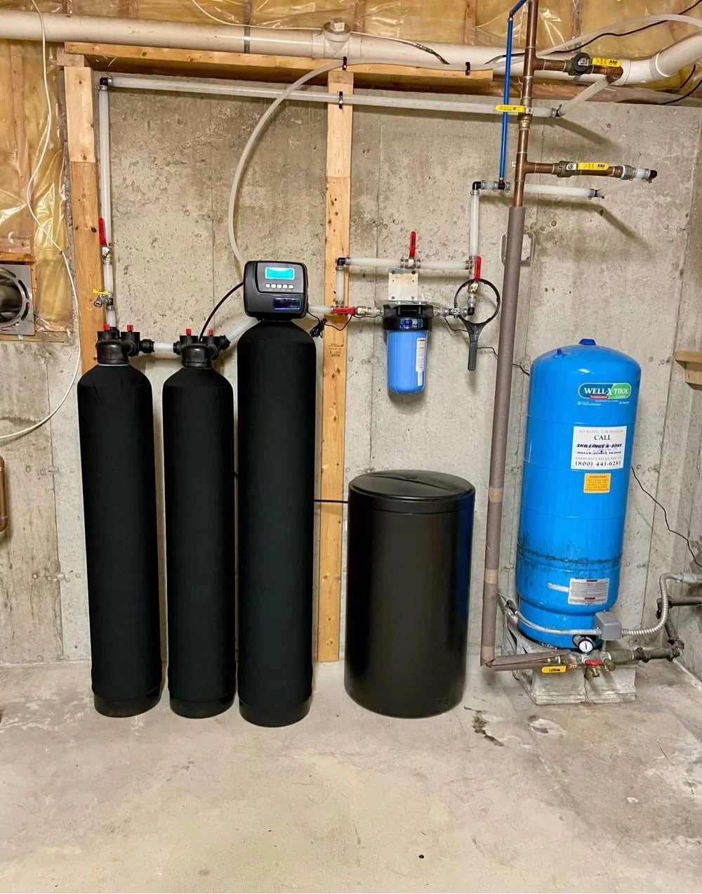 A residential water filtration system installed on a concrete wall with pipes, valves, a black softener tank, a blue pressure tank, and black filter tanks.