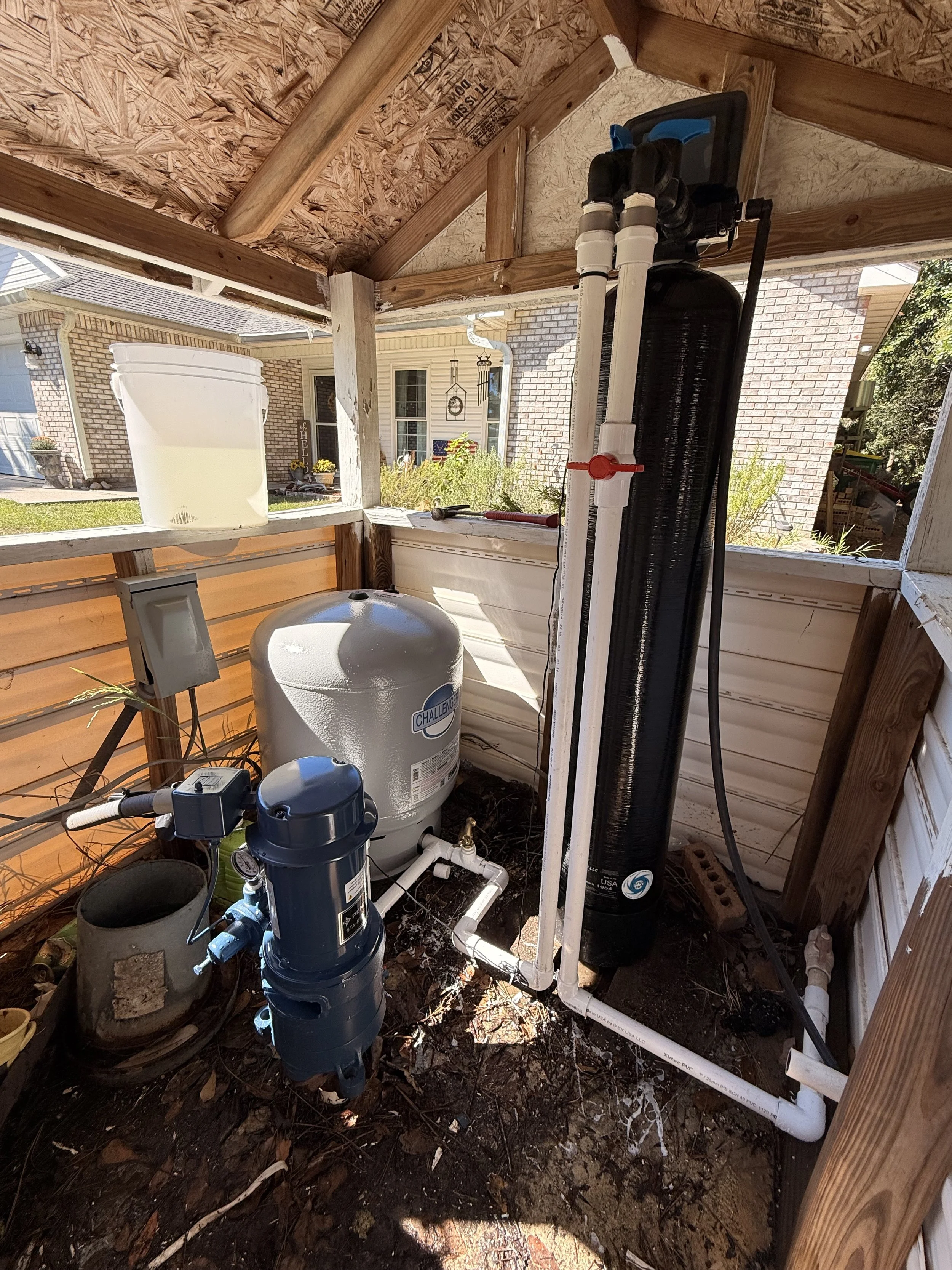 Setup of a home water filtration system on a porch, including a large black reverse osmosis tank, a blue pump, white PVC pipes, a pressure tank, and a bucket, with brick and siding exterior background.