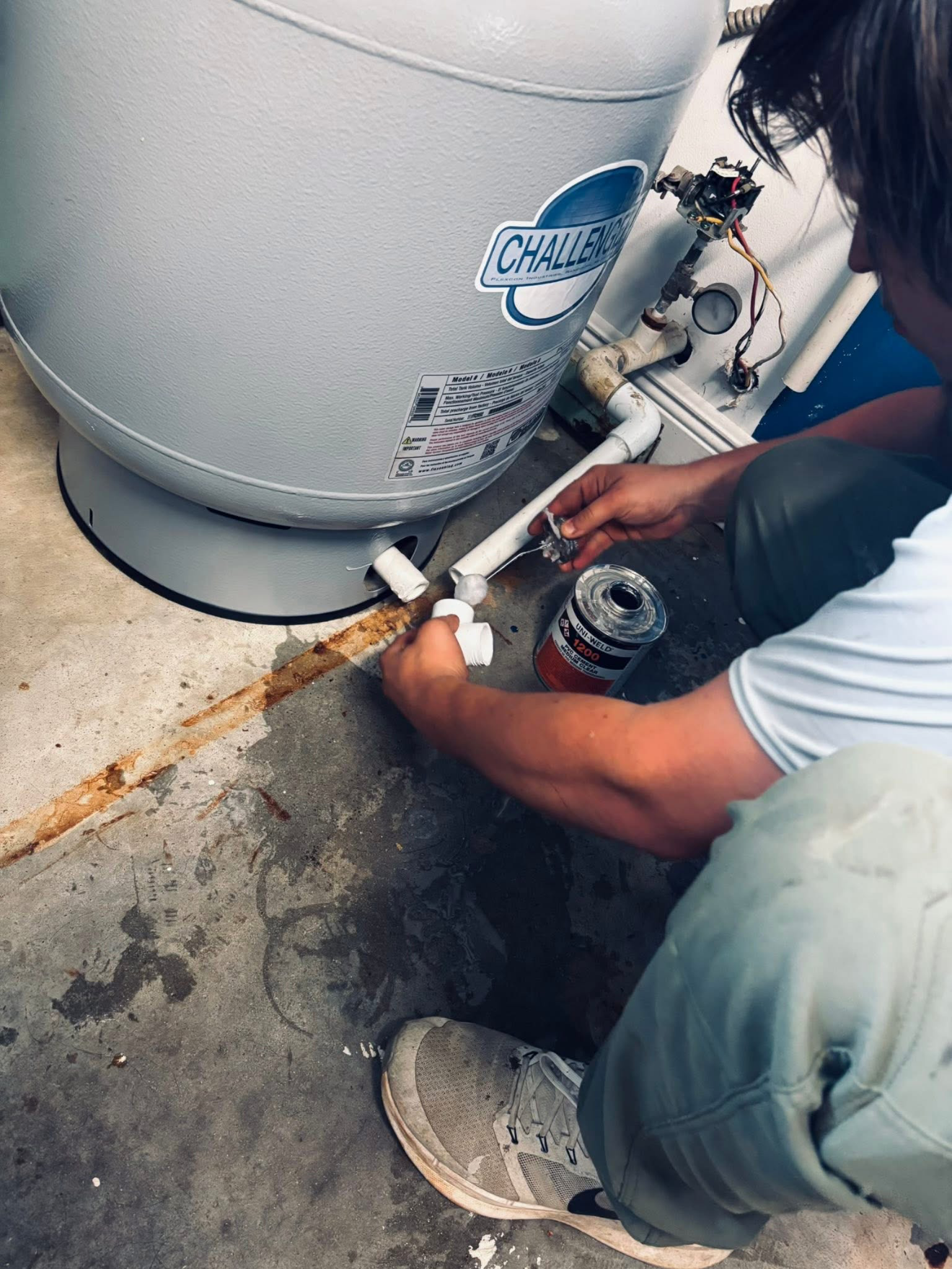Person working on the plumbing of a hot water heater, connecting pipes on a concrete floor.