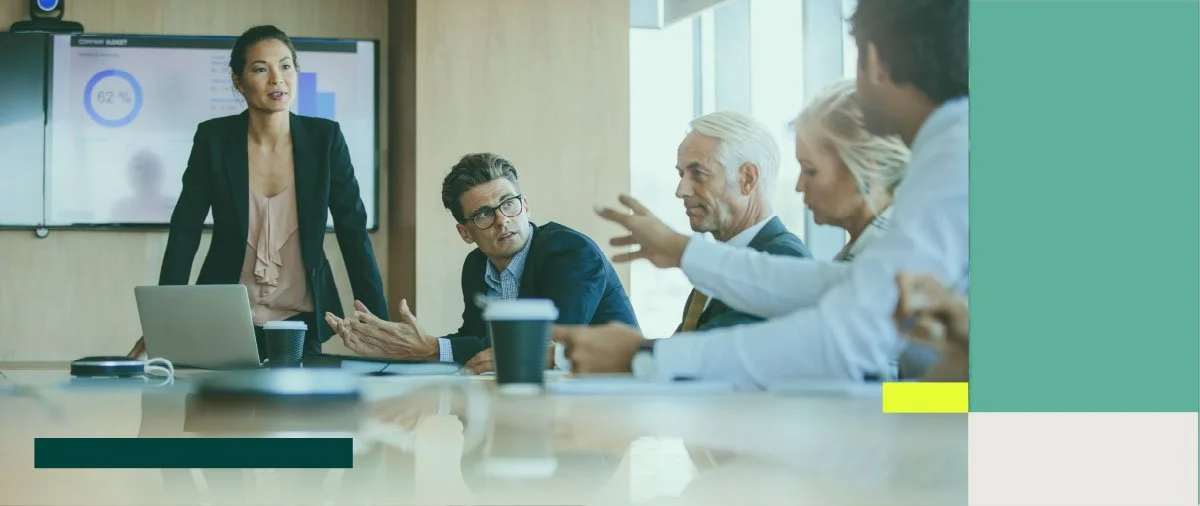 Franchisor business meeting with five professionals in a conference room, one woman standing near a laptop and screen, four seated and engaged in discussion.