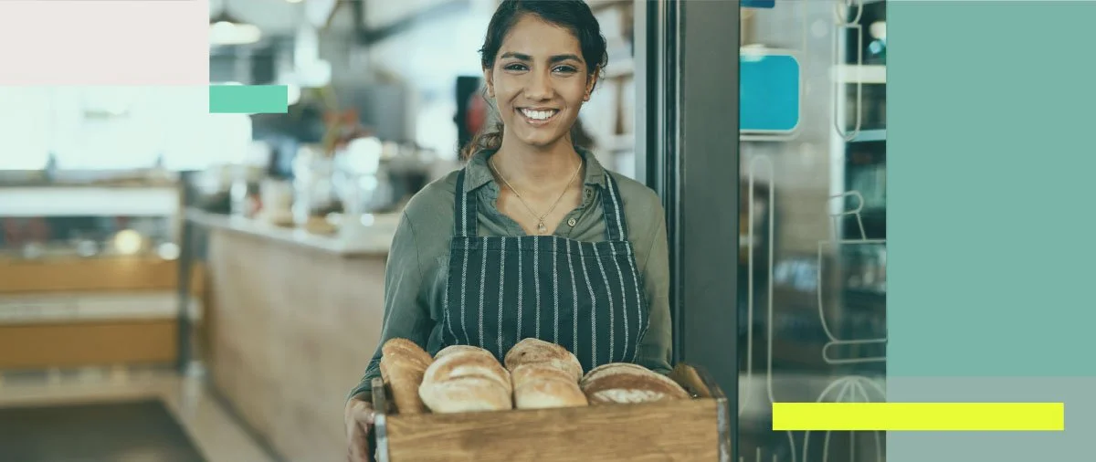 Smiling woman in apron holding a tray of baked bread in a franchise bakery.