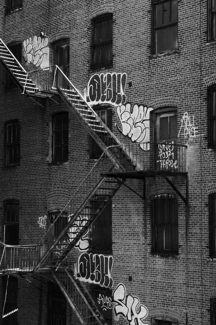 Black and white photo of an old brick building with graffiti art on the wall. There is a metal fire escape zigzagging across the building.