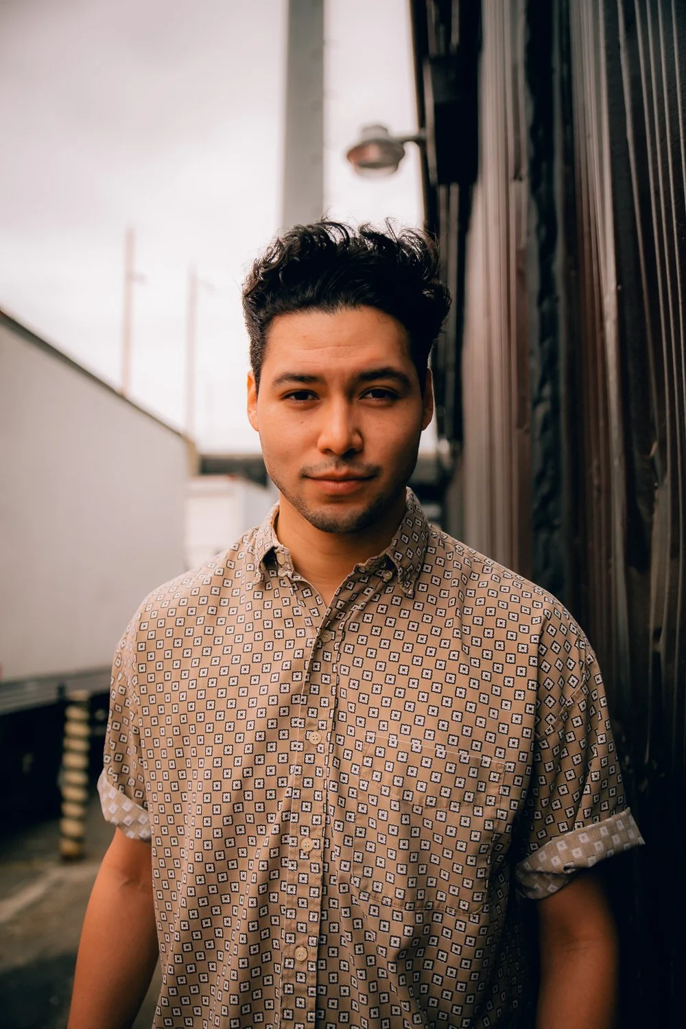 A young man with dark, curly hair and a light brown patterned short-sleeve shirt poses in an outdoor urban setting with containers and a streetlamp behind him.