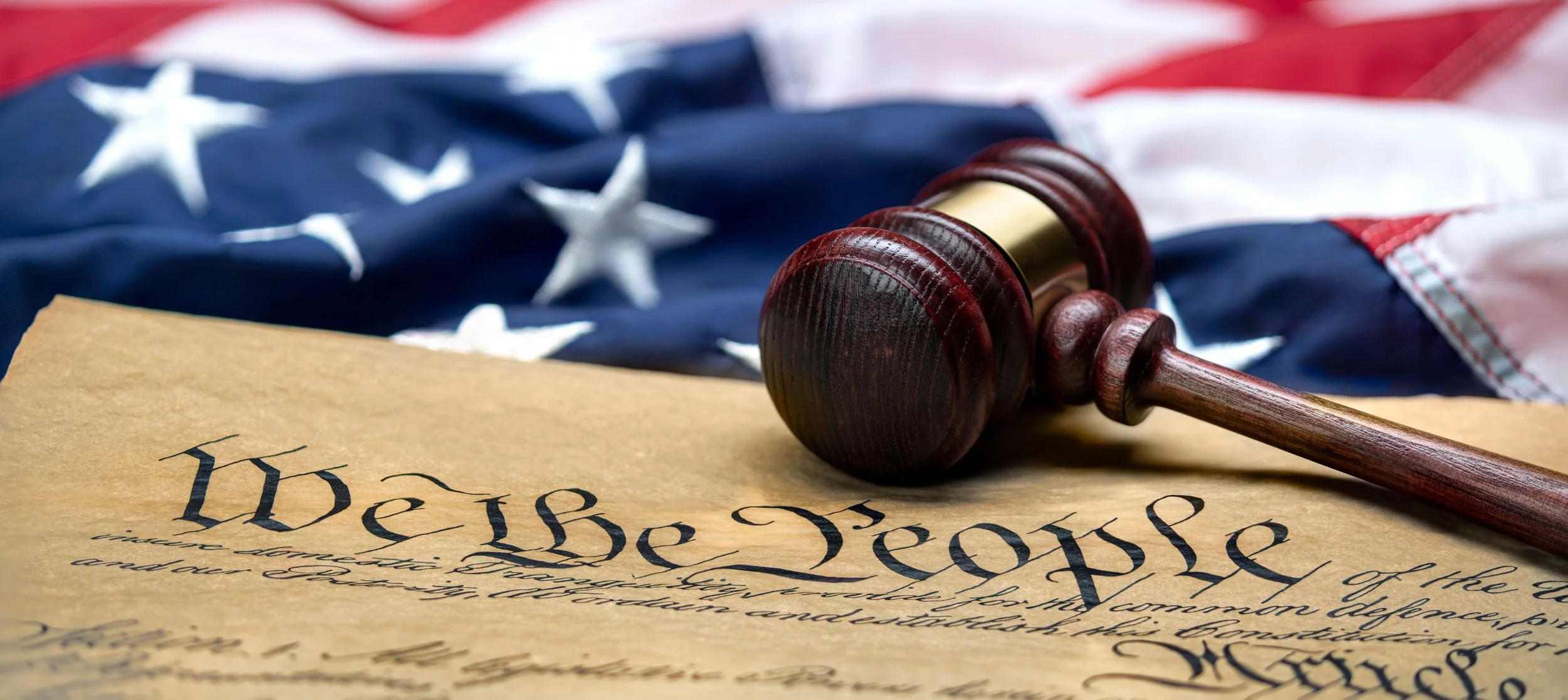 A brown, mostly wood gavel and a corner of a copy of the Declaration of Independence with words “We The People” lie on top of an American flag that is only partially shown. Tags: Justice, Democracy & Resistance.