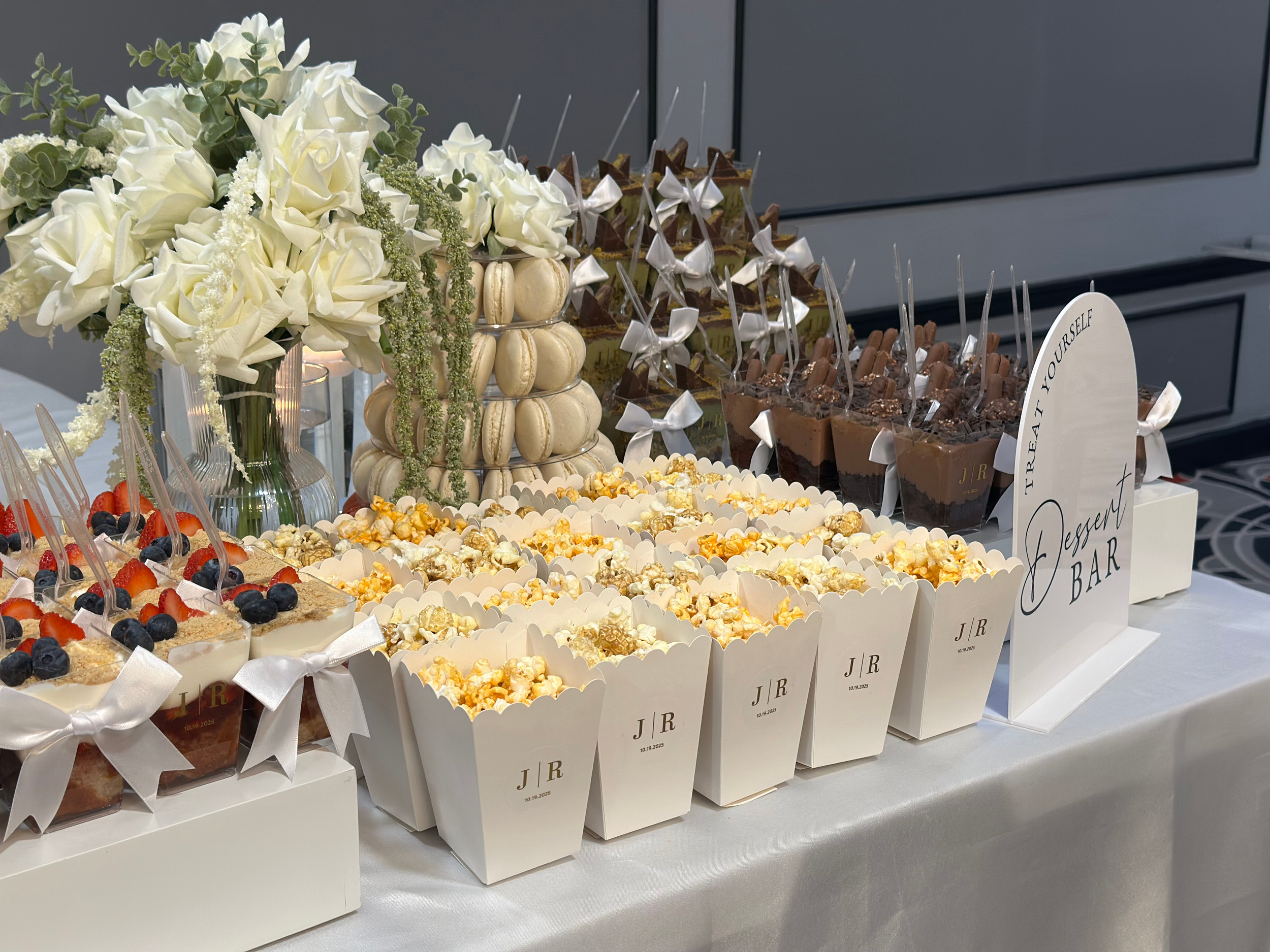 An assortment of desserts on a table with white flowers. Includes fruit-topped cakes, popcorn boxes, macarons, and chocolate mousse cups, all decorated with ribbons and labels.