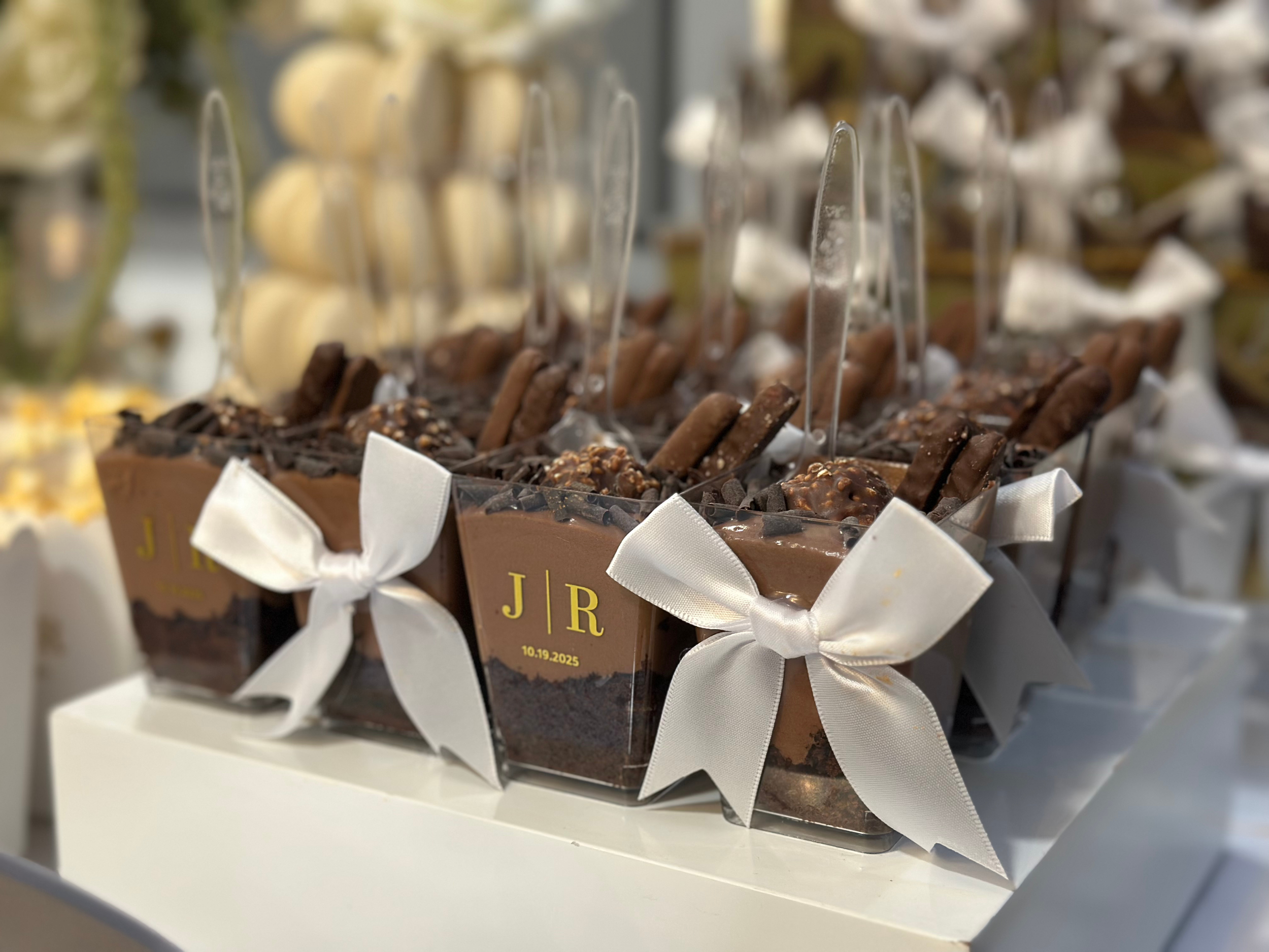 Decorative chocolate dessert cups with white ribbons, topped with chocolate sticks and various chocolates, labeled with initials 'J R' and a date '10.19.2025' at a celebration or wedding event.