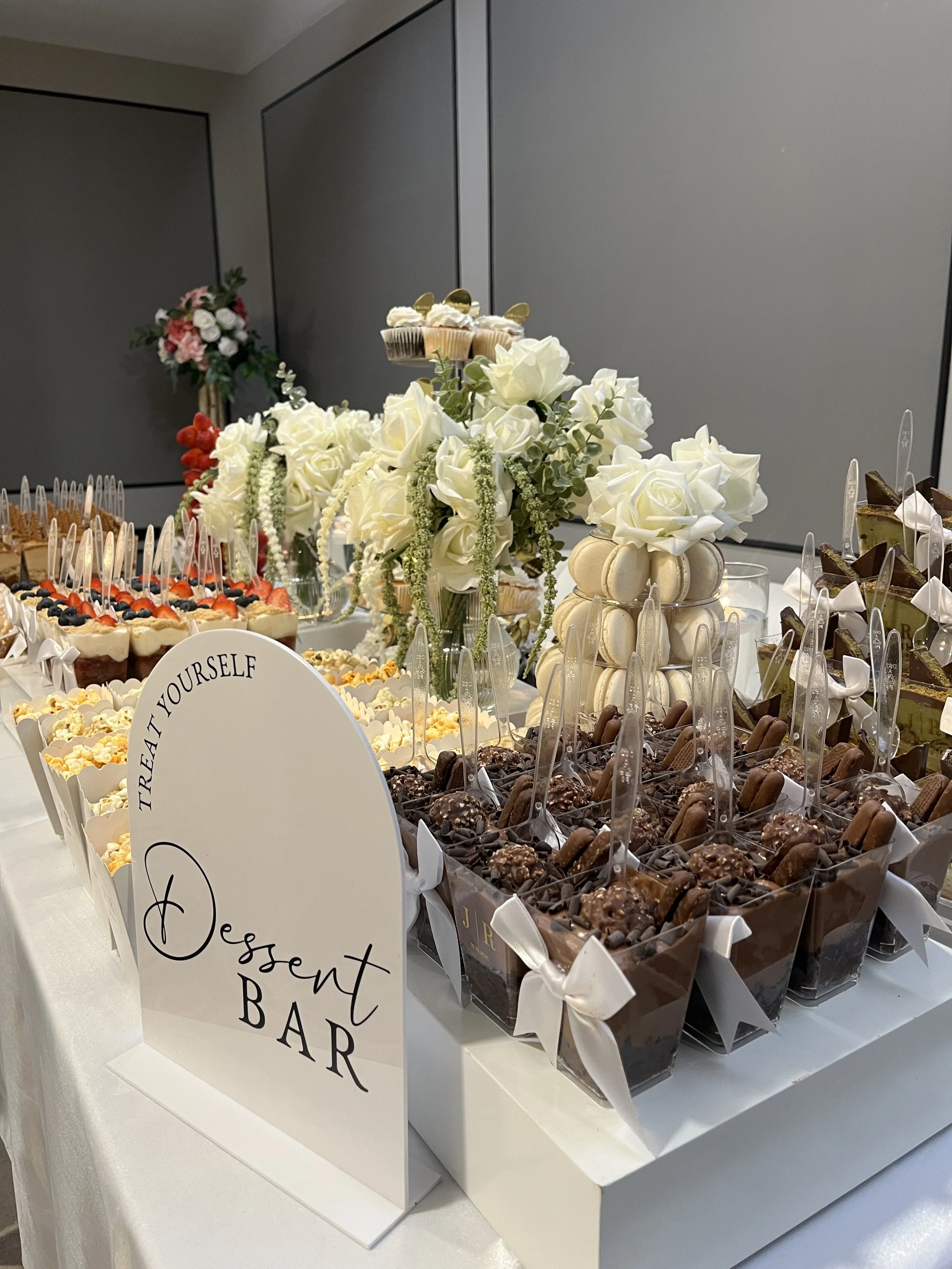 A dessert table at an event displays various treats, including chocolates, cupcakes, and berries, with a centerpiece of white roses and greenery. A sign reads 'Treat Yourself Dessert Bar'.