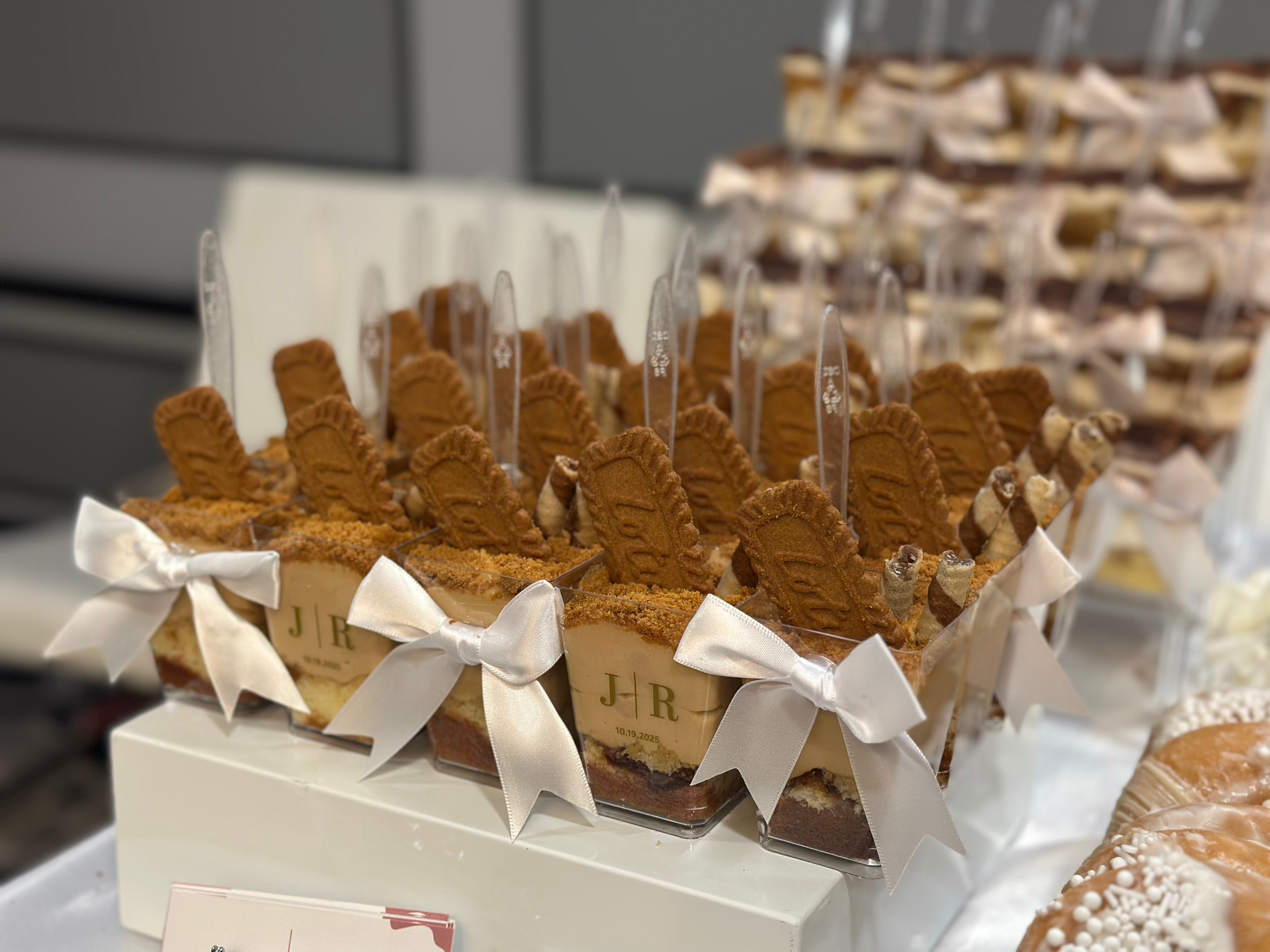 A display of individual dessert cups decorated with cookies, ribbons, and small plaques, with a background of more desserts.