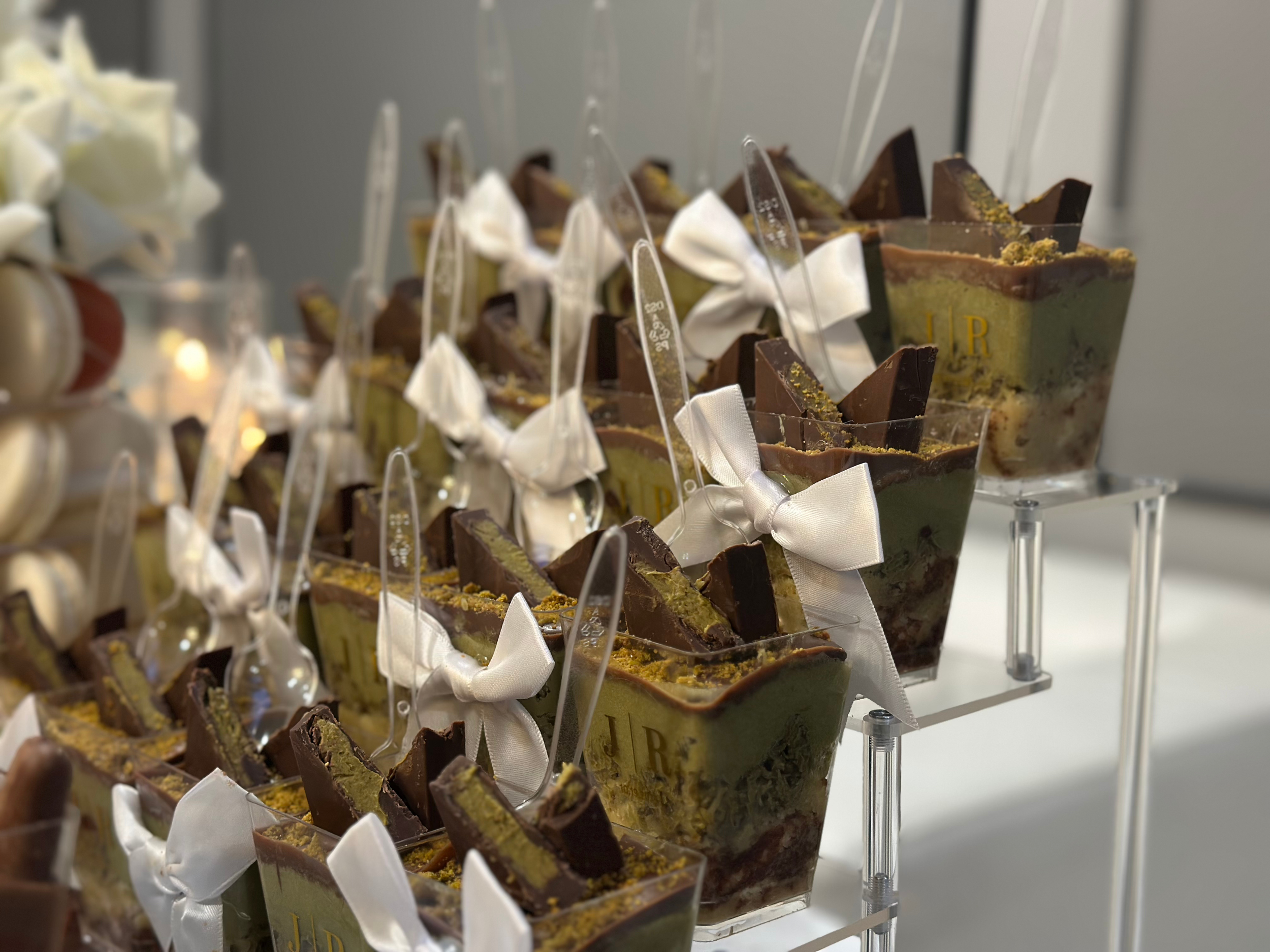 Individual dessert cups filled with layered mousse and topped with chocolate pieces, white bows, and decorative clear sticks, arranged on a clear display stand at an event.