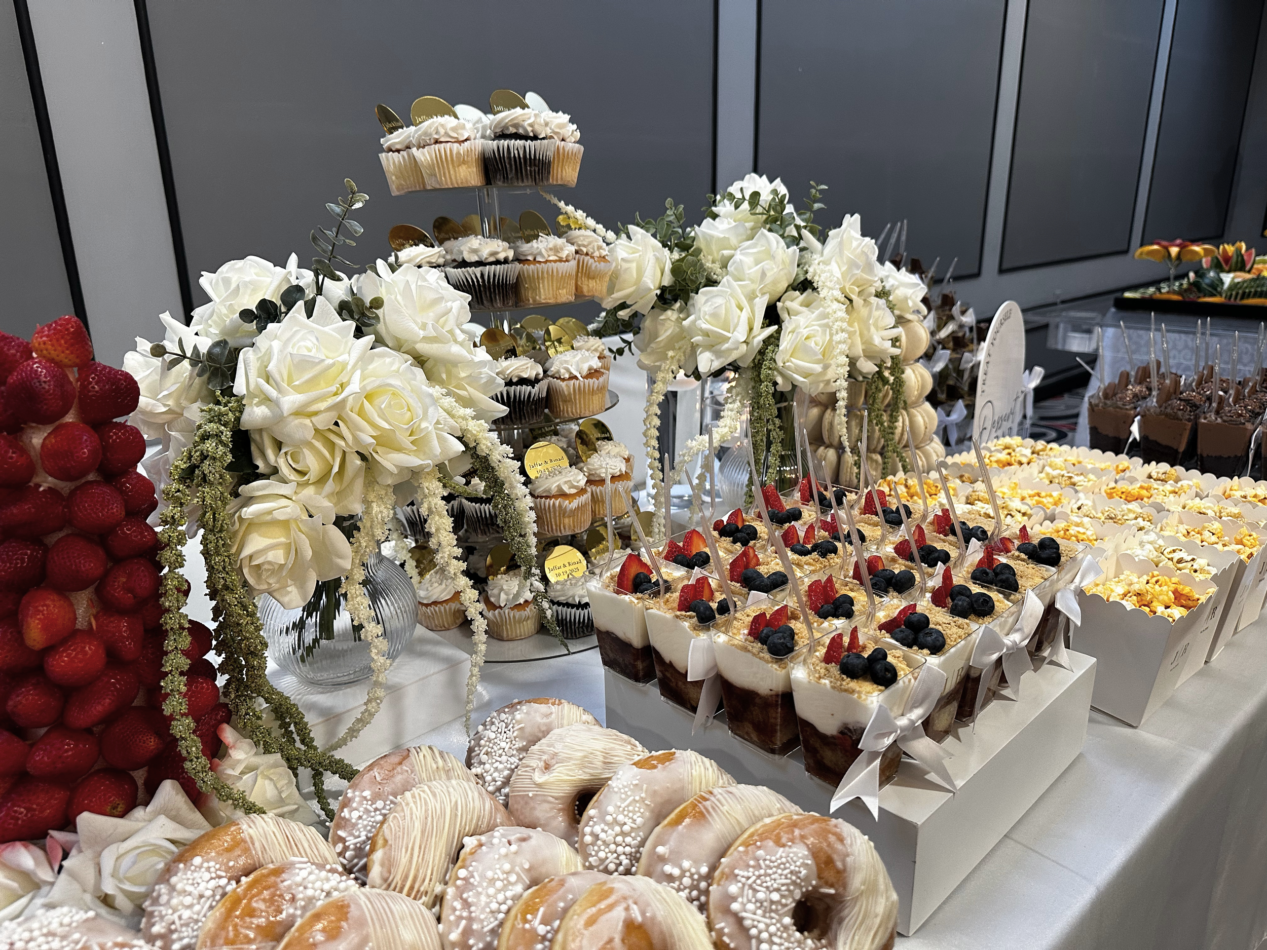 A dessert table with strawberries, cupcakes, parfaits topped with berries, and donuts decorated with icing and sprinkles, all decorated with white flowers and greenery.