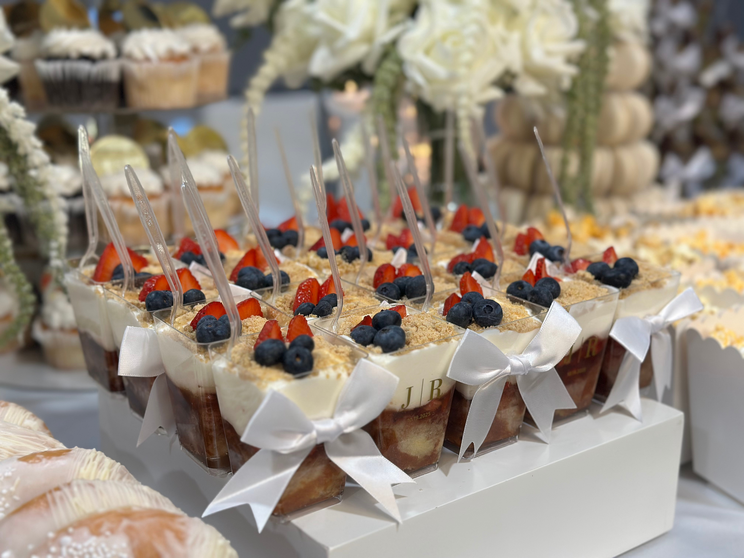 Small dessert cups with whipped cream, strawberries, blueberries, and crumb topping, decorated with white ribbons, displayed on a white stand, with additional desserts in the background.
