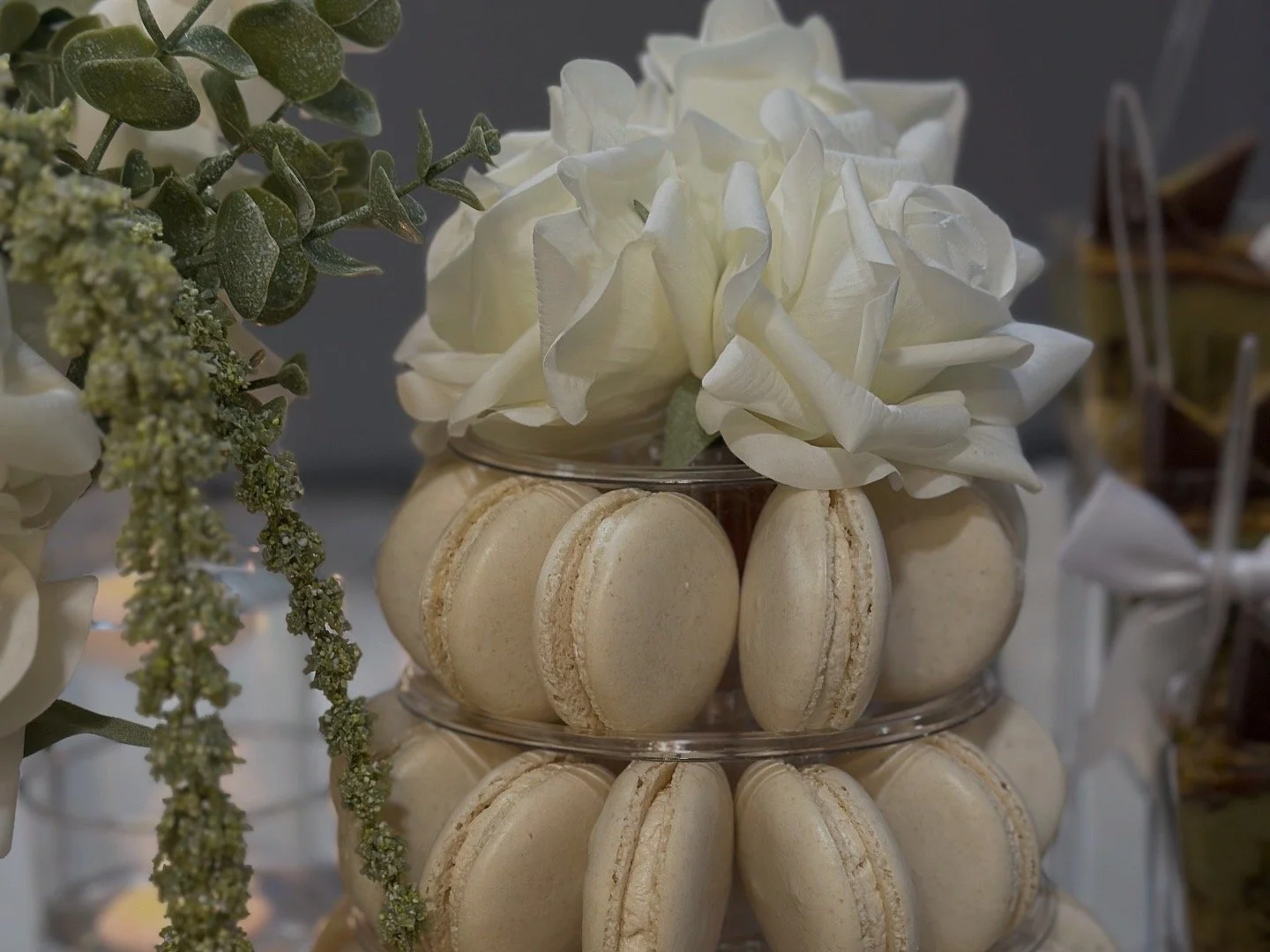 A tiered display of cream-colored macarons with white flowers on top, surrounded by green foliage.