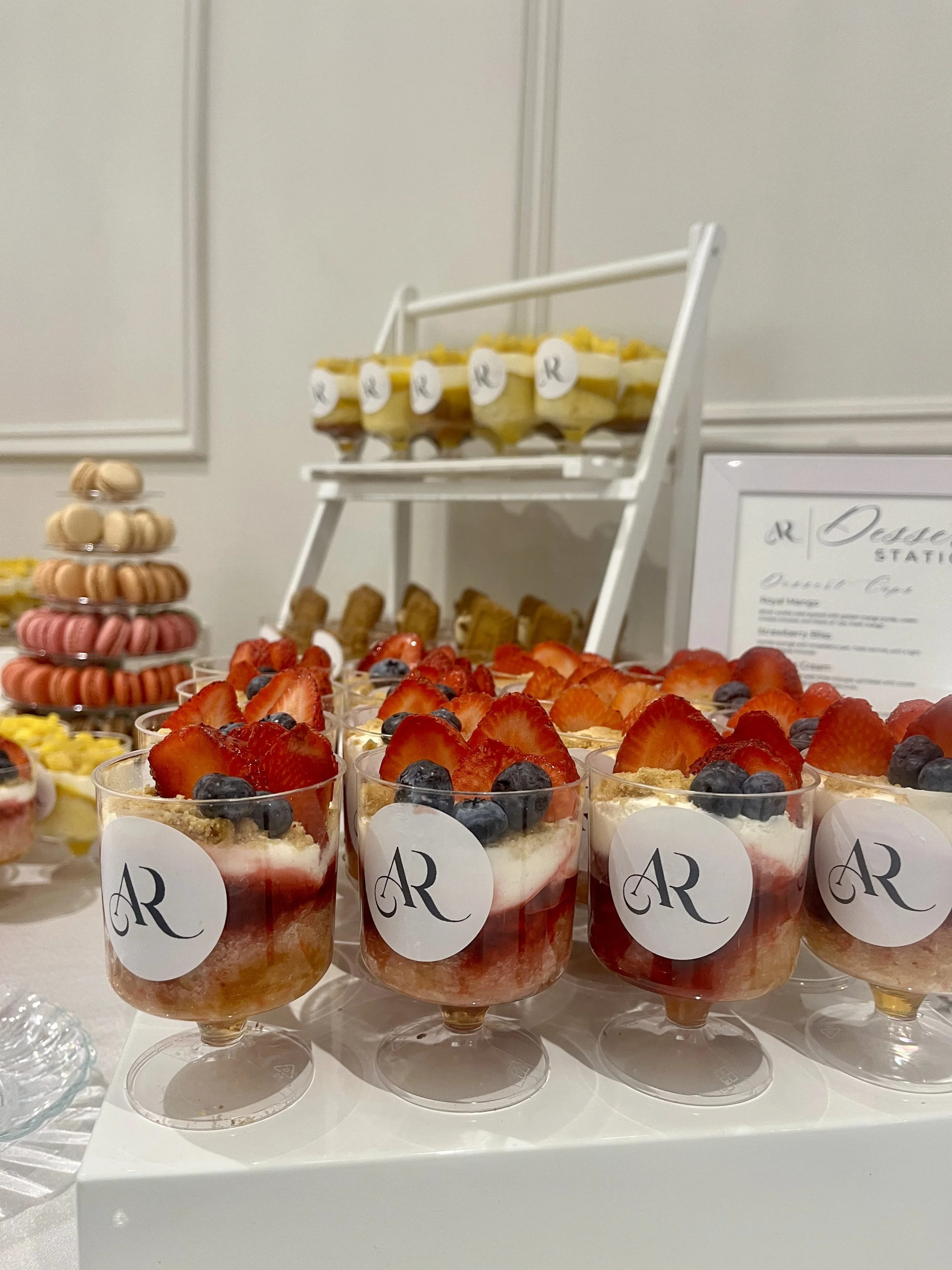 Dessert table featuring individual strawberry and blueberry parfaits with white AR logos, layered cakes with fruit toppings, and a macaron display on a white table.