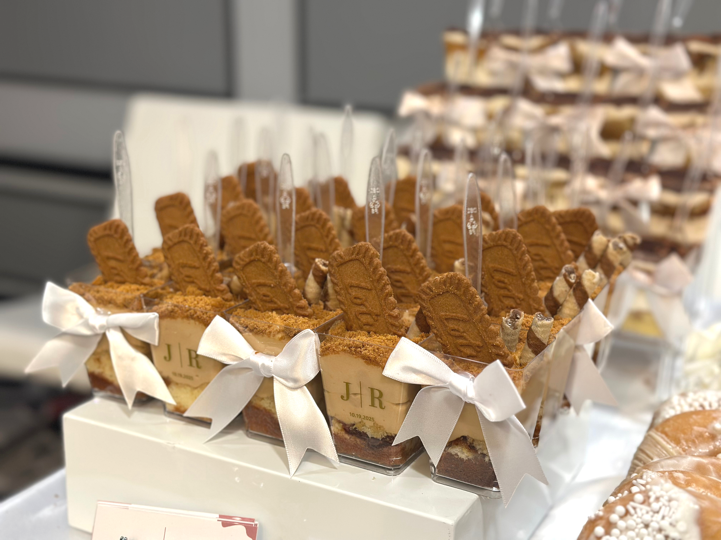 A decorated dessert display with individual parfait cups topped with Biscoff cookies, white ribbons, and teaspoons. In the background, there are more desserts and a small sign with initials and a date.