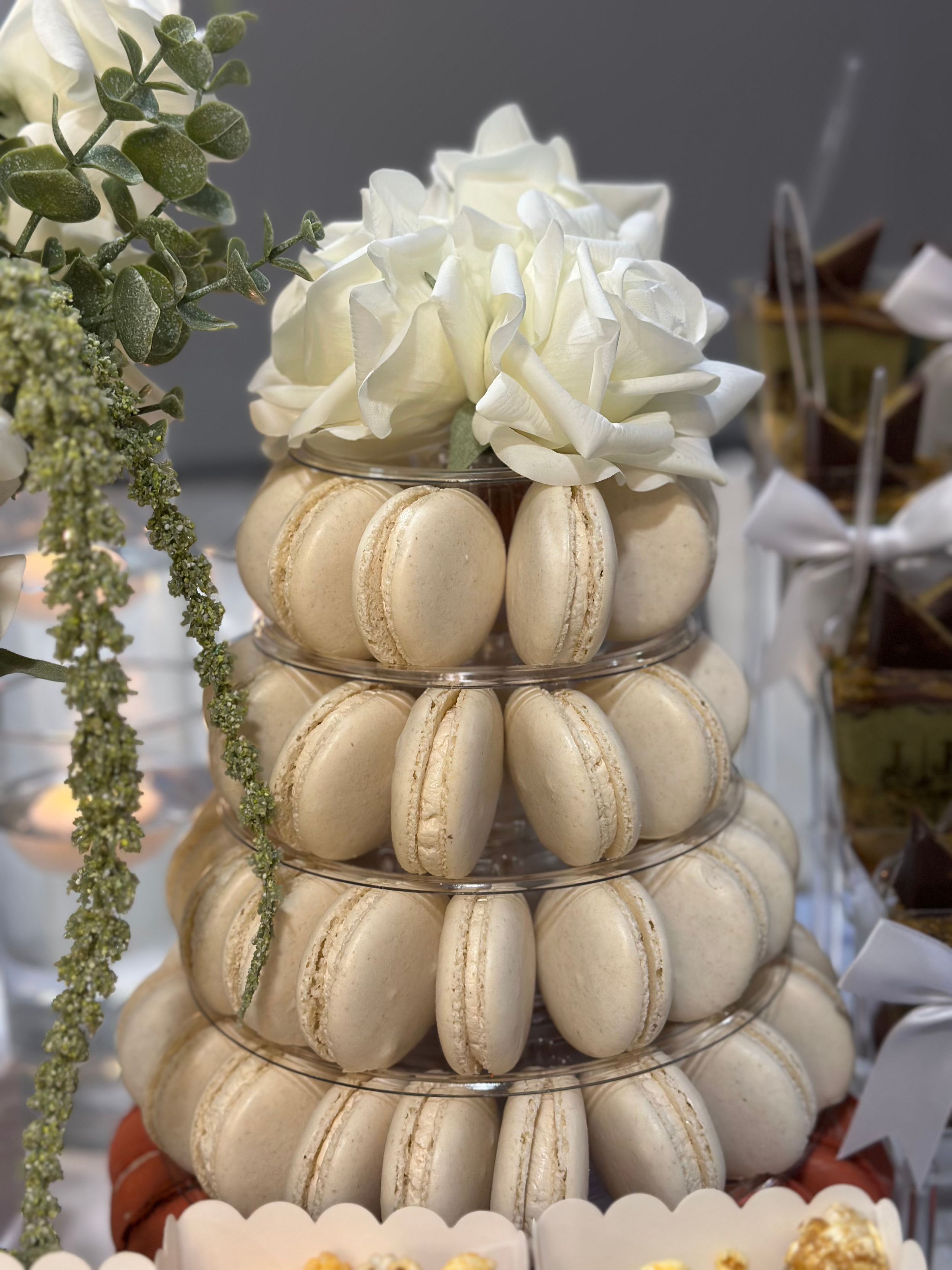 A multi-tiered display of white macarons topped with white roses and greenery, arranged in a clear circular stand on a table.