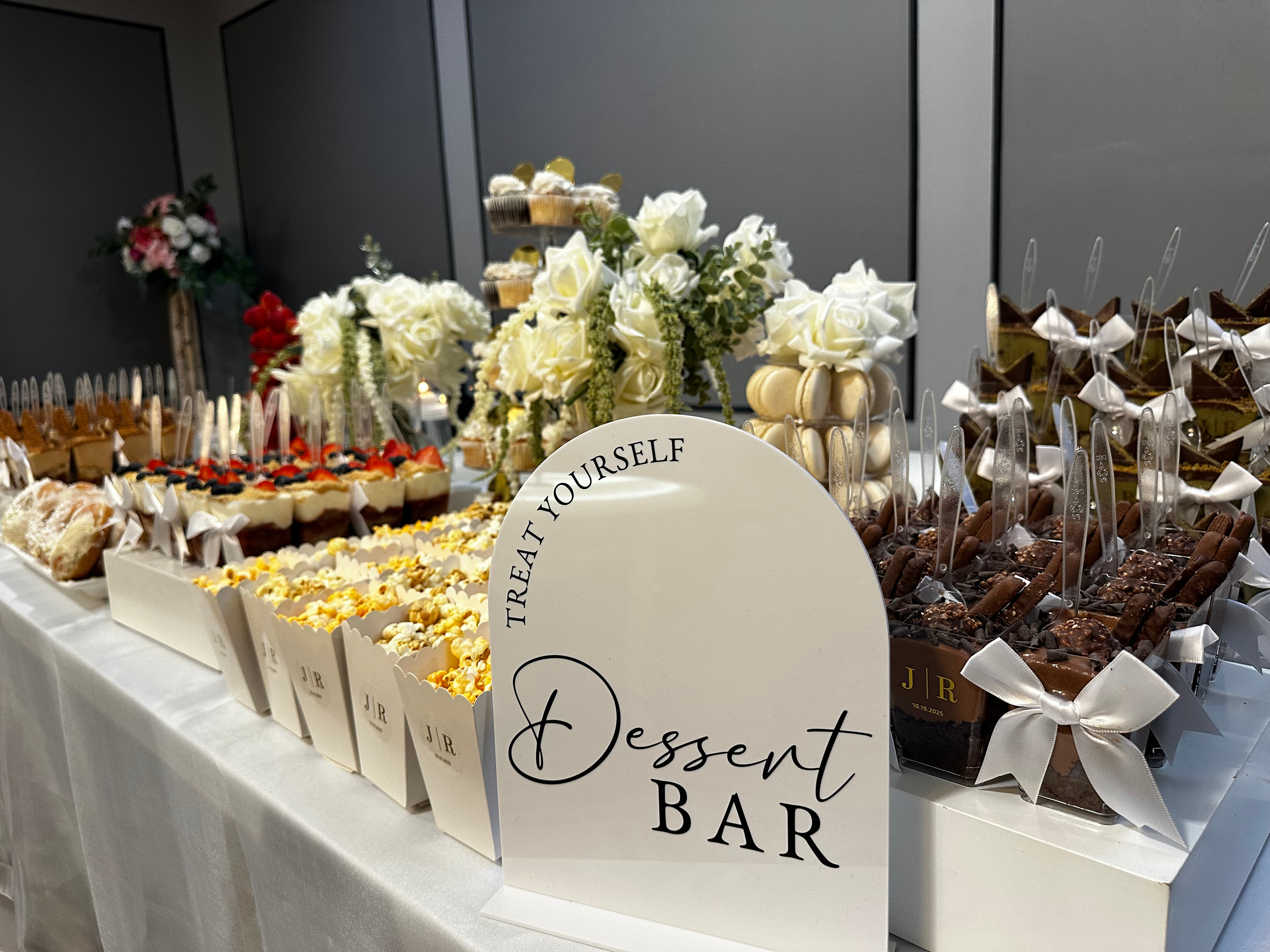 A dessert bar with an assortment of sweet treats including cupcakes, cookies, and chocolates, decorated with white flowers and a sign that reads and 'Treat Yourself Dessert Bar'