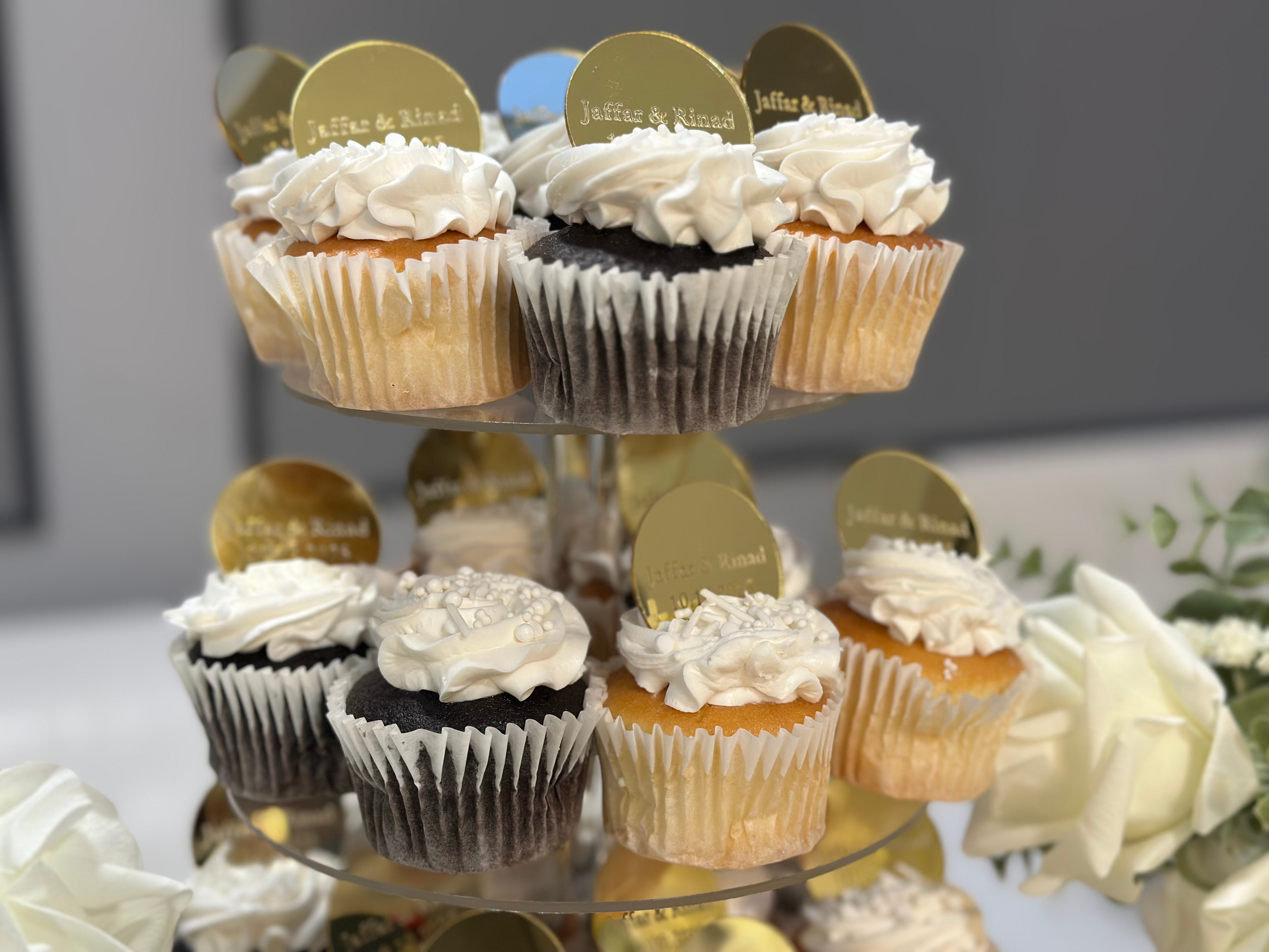 Assorted cupcakes on a two-tiered stand, decorated with white and dark frosting, some with gold edible decorations
