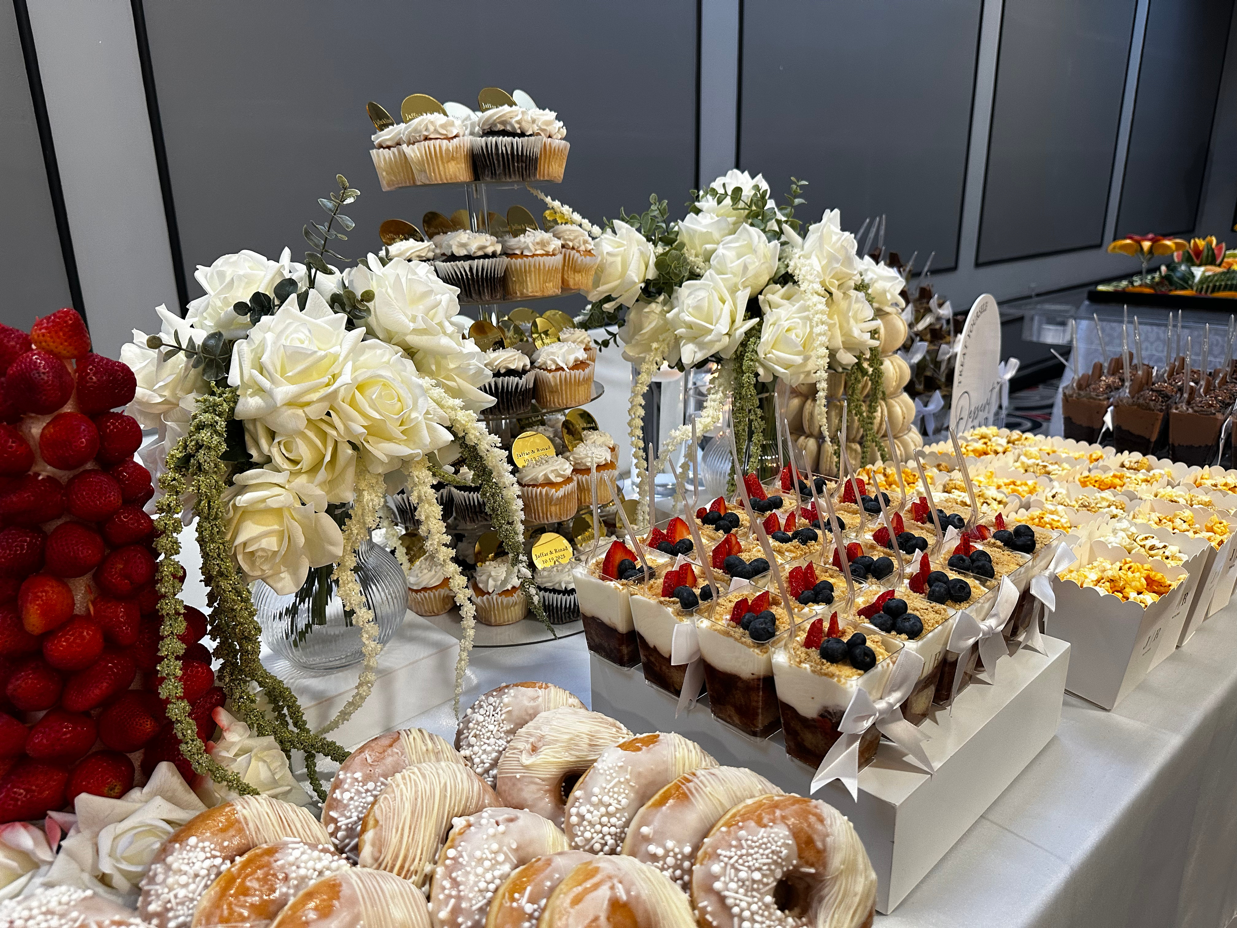 Dessert table with cupcakes, berry parfaits, donuts, and strawberries, decorated with white roses and green foliage.