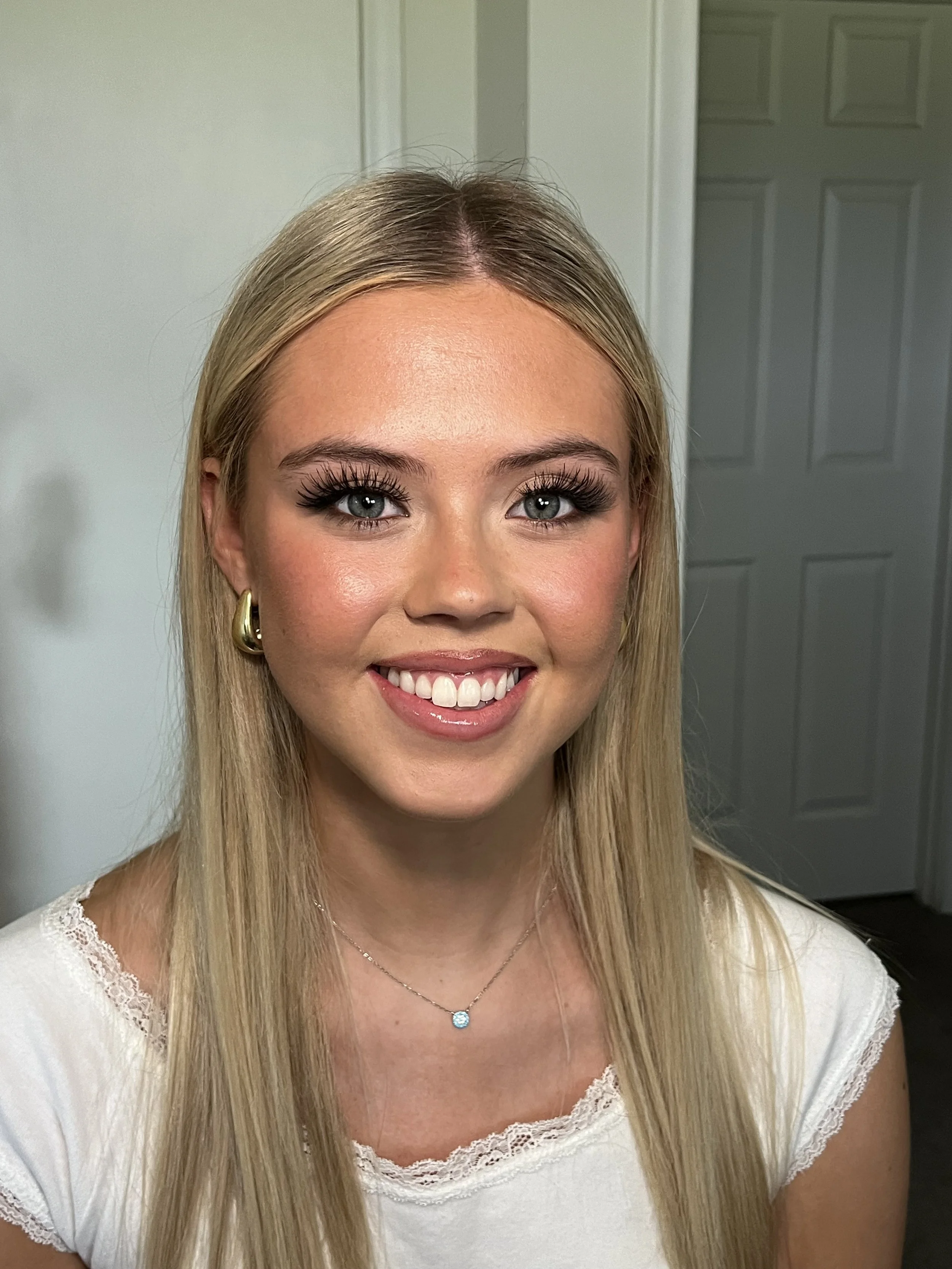 Close-up of young woman with long blonde hair, gray eyes, wearing gold hoop earrings and a small necklace, smiling with white teeth, inside a room with white walls and a closed white door in the background.