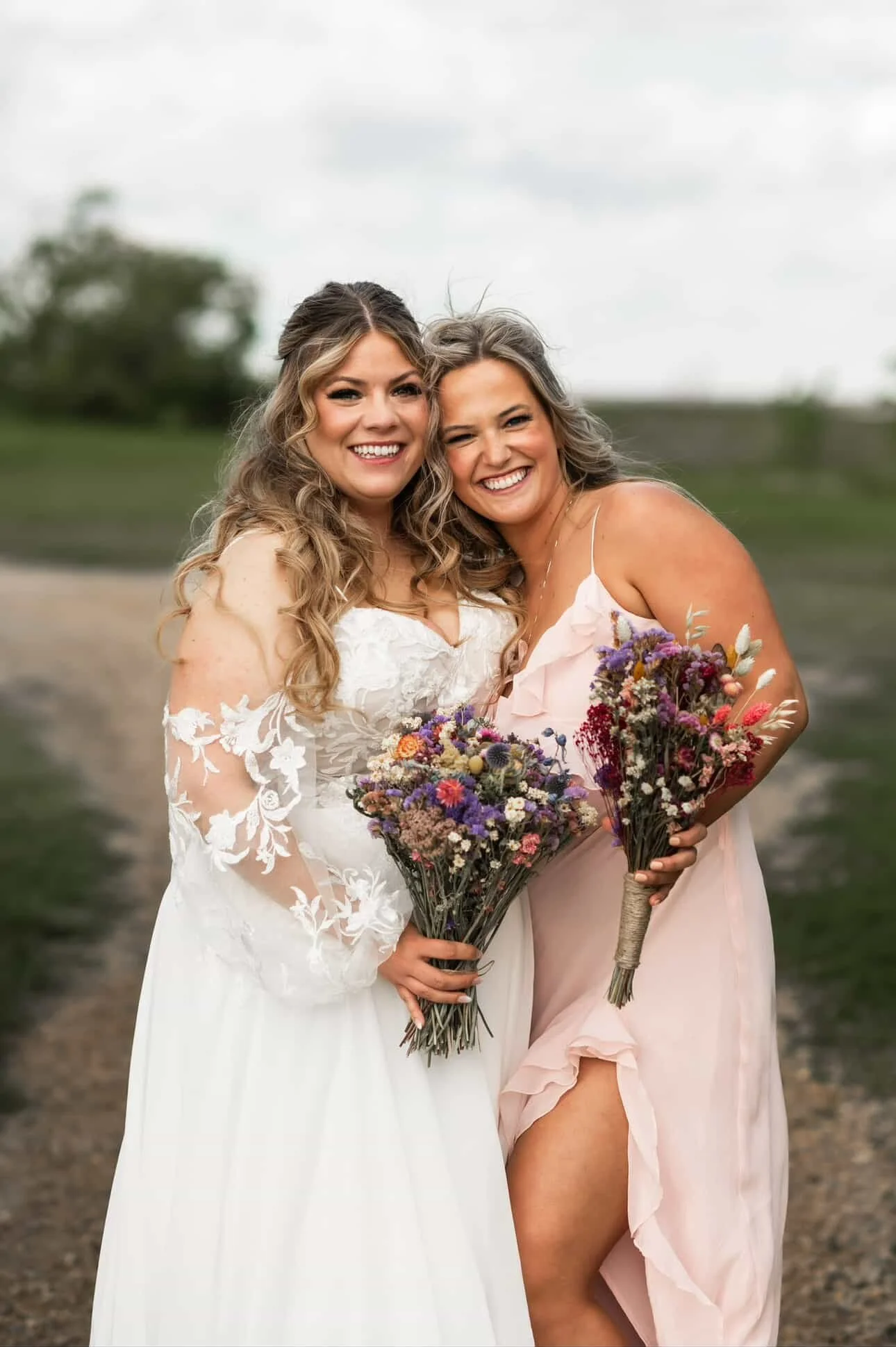 Two women standing outdoors, smiling, one in a white wedding dress and the other in a light pink dress, holding bouquets of flowers.
