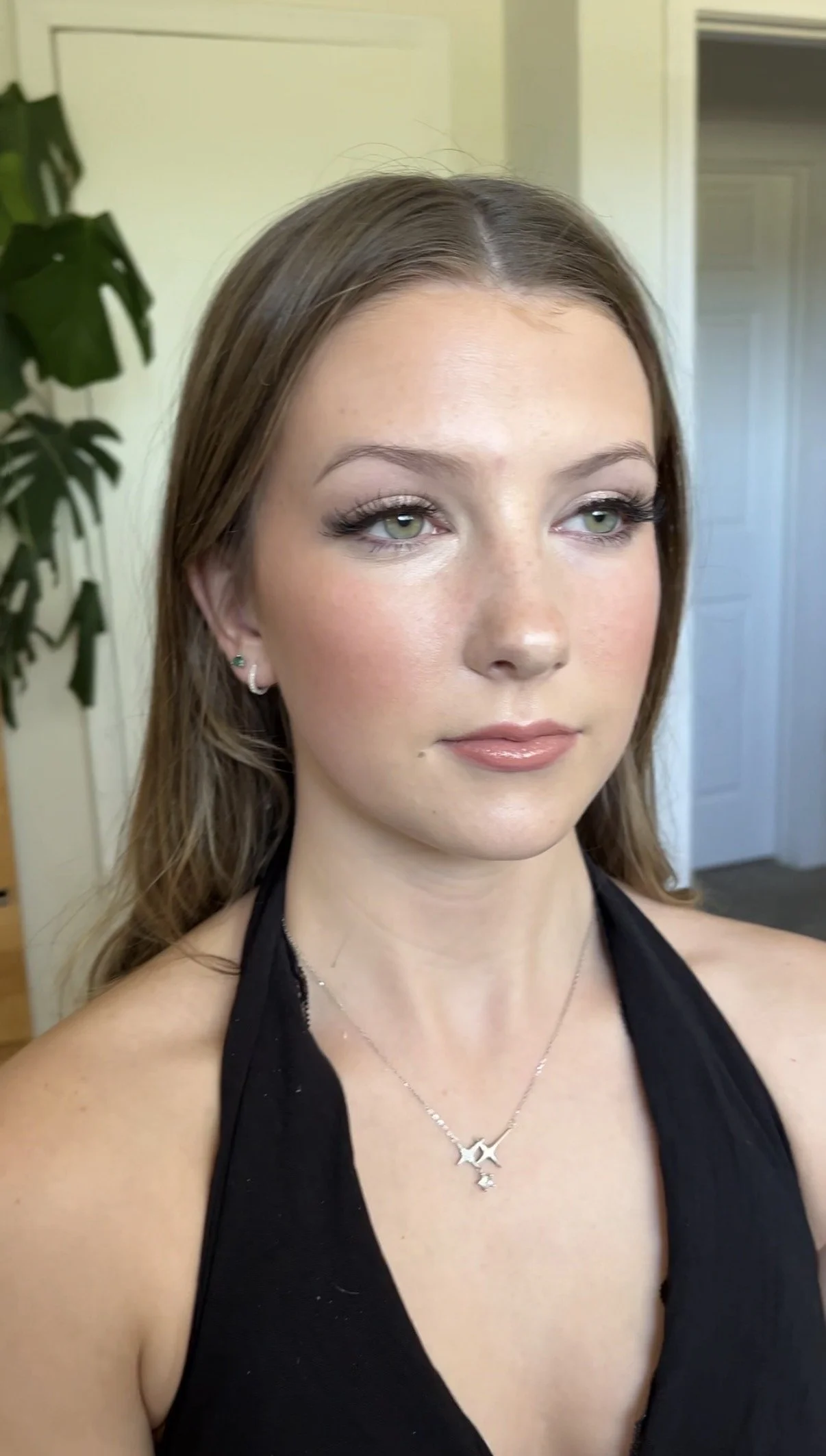 A young woman with long, wavy brown hair and fair skin, wearing eye makeup and glossy lips, is dressed in a black halter top and silver jewelry, including a star-shaped necklace and earrings, standing indoors near a white door and a green plant.