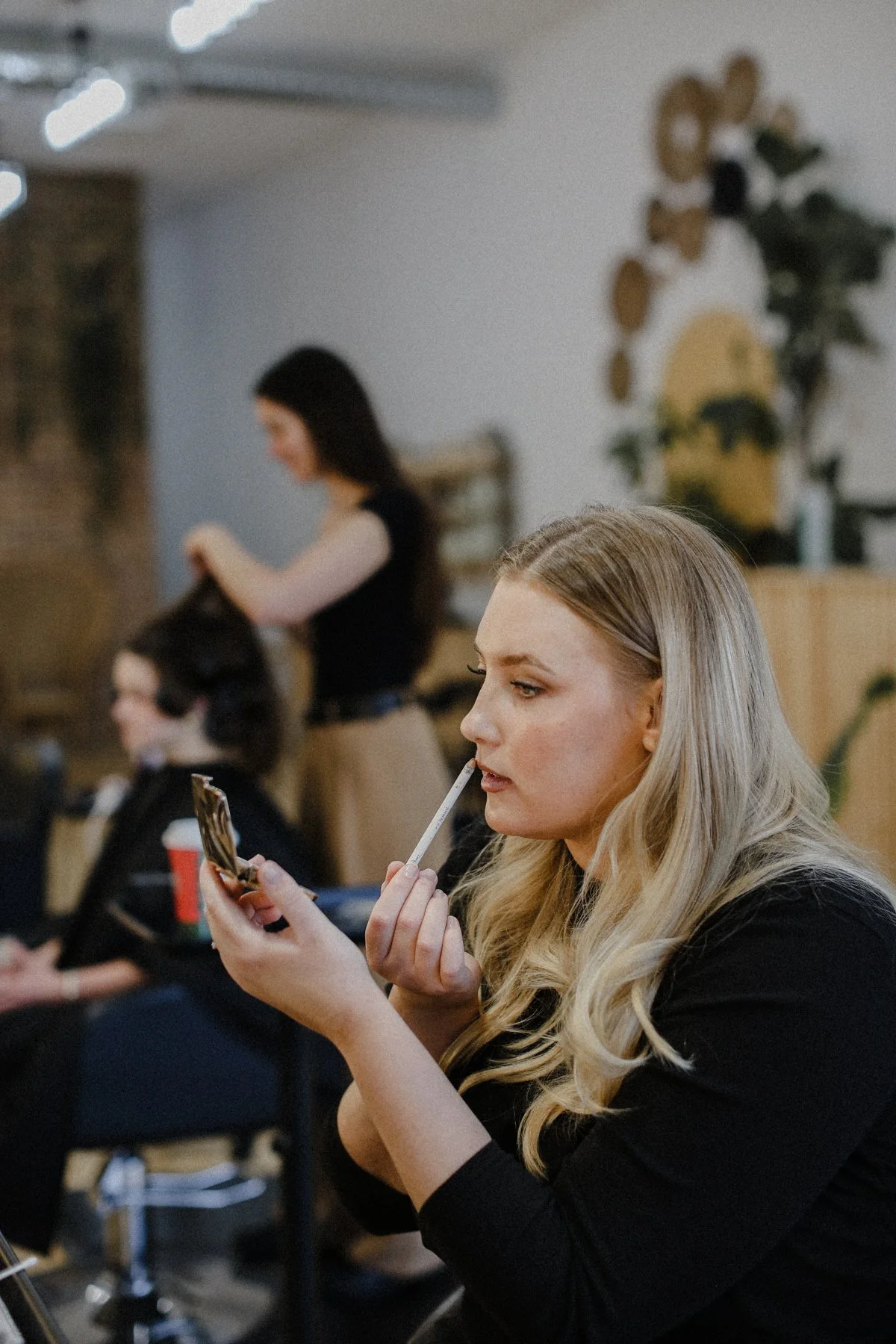 A woman with long blonde hair applying makeup while looking into her compact mirror in a salon or barbershop.