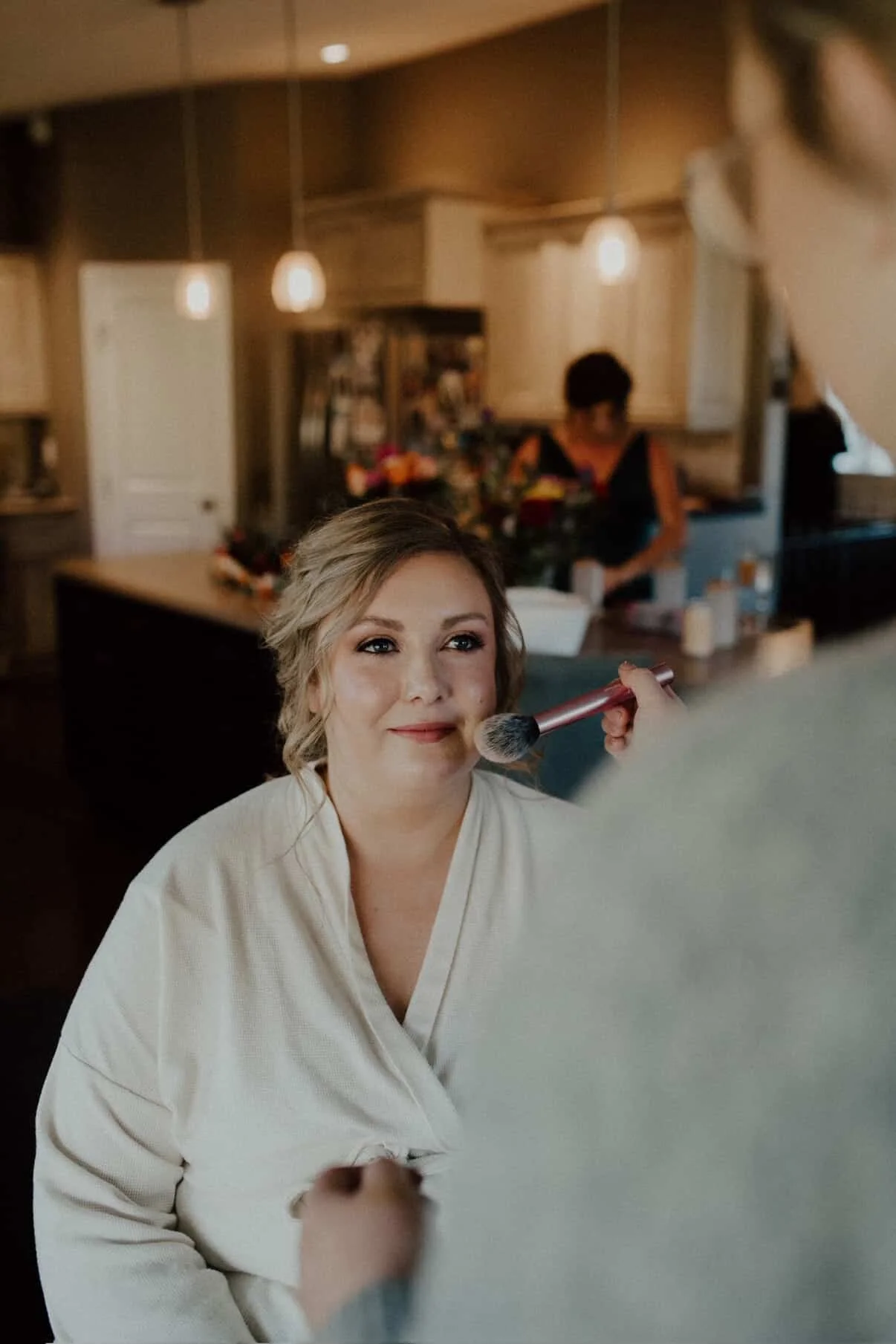 Woman with blonde hair receives makeup application using a brush in her cheek in a cozy home setting.