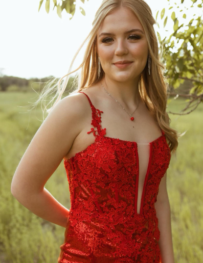 Young woman in a red lace dress outdoors, standing near a tree with a field in the background, smiling at the camera.