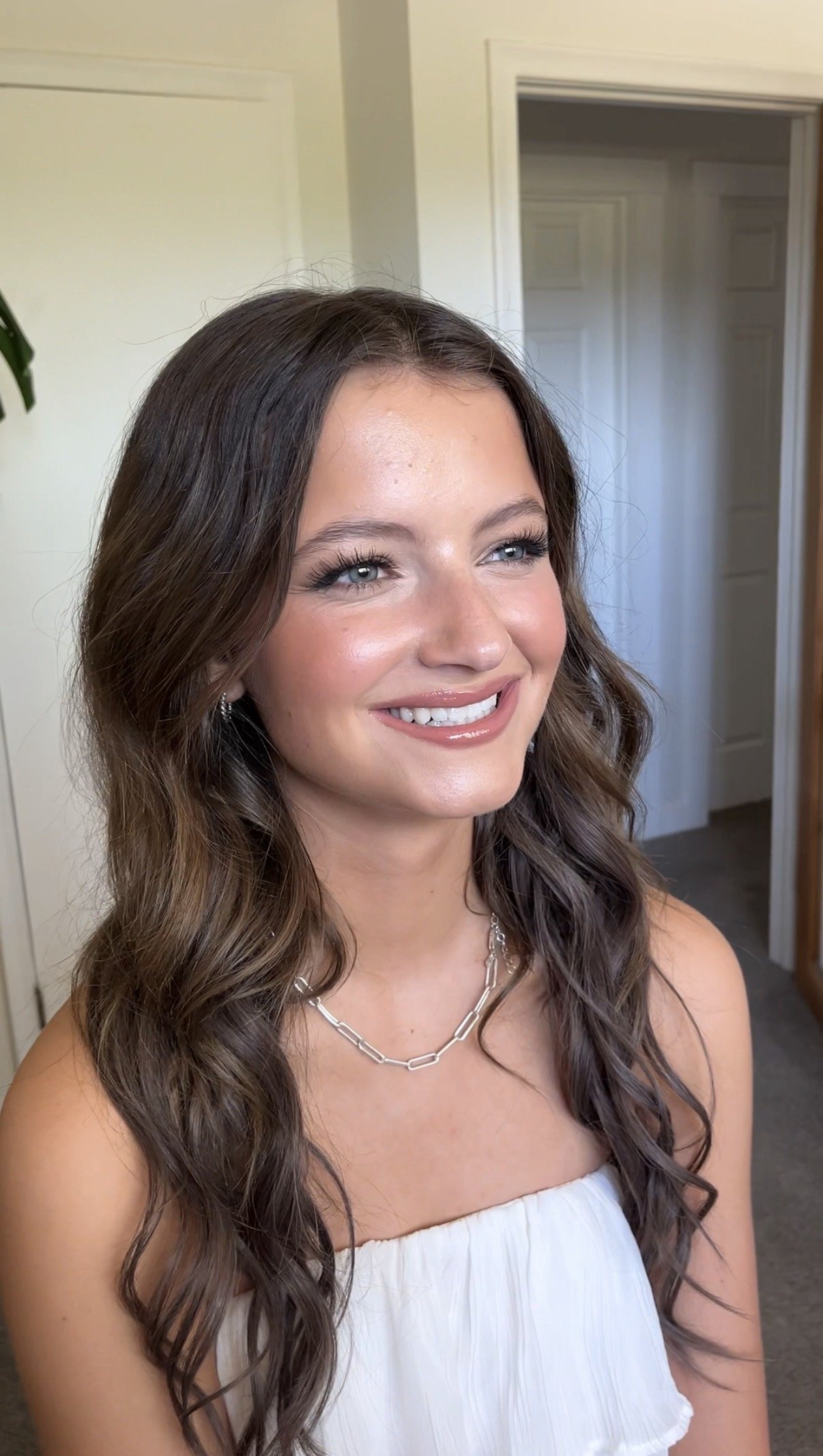 A young woman with long wavy brown hair, light skin, and blue eyes, smiling in a white strapless dress with a silver chain necklace, standing indoors near a door.