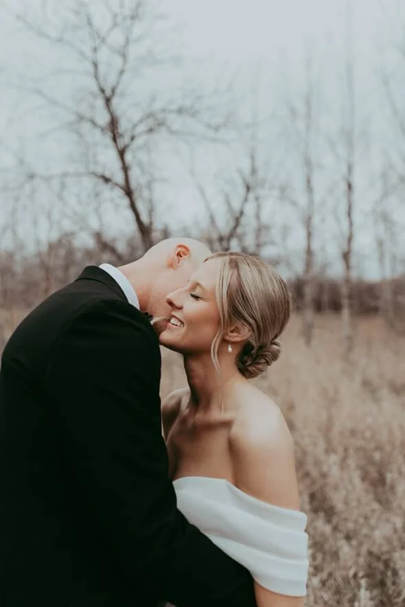A bride and groom smiling and embracing outdoors during their wedding, with leafless trees in the background.
