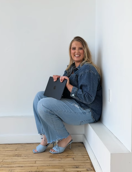 Young woman sitting on a built-in white bench in a minimalist room, holding a closed laptop, smiling, wearing a denim jacket, jeans, and light blue shoes.