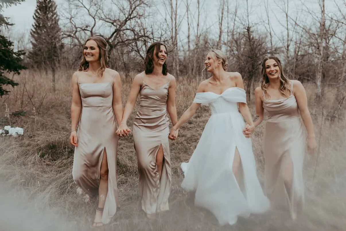 Four women dressed in formal gowns, holding hands, walking outdoors in a natural setting with leafless trees, smiling and enjoying each other's company.