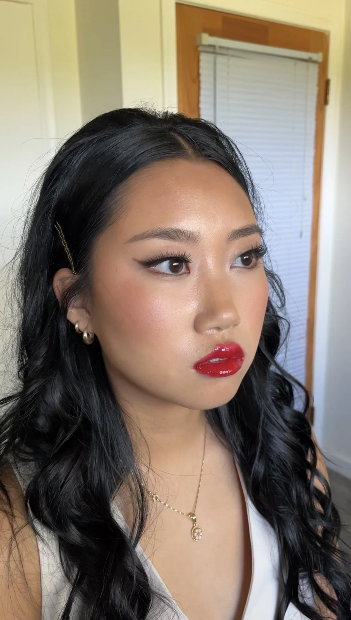 Close-up of a woman with long black wavy hair, wearing gold hoop earrings and a delicate gold necklace with a pendant, with makeup featuring red lipstick, eyeliner, and contoured cheeks, in front of a wooden door and white wall.