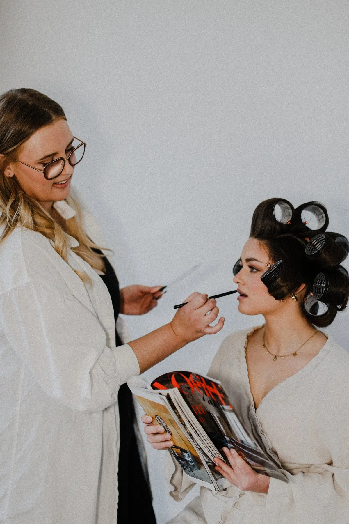 A woman getting her makeup done with hair rollers while holding a magazine