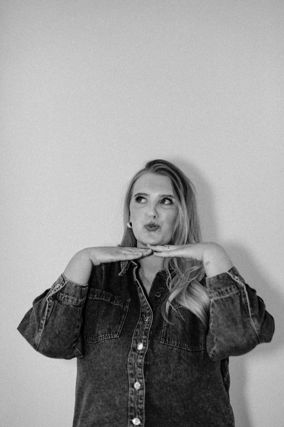 A woman with long hair, wearing a denim shirt, poses with her hands under her chin and a playful expression, looking upward.
