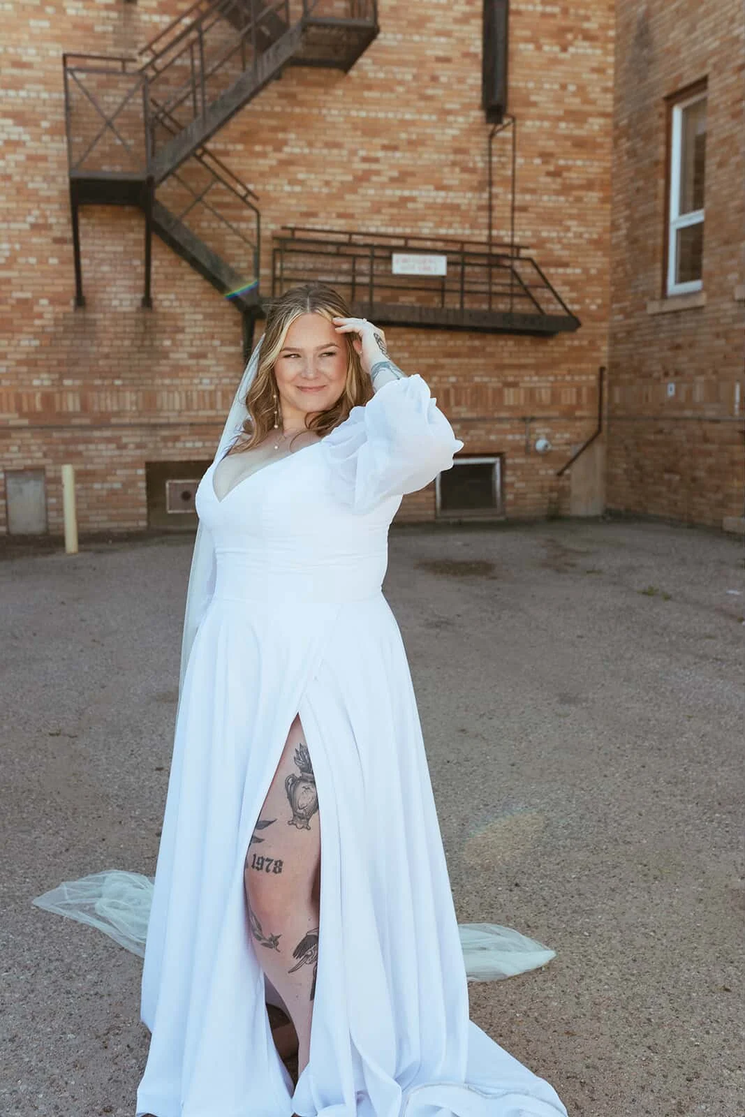 A woman in a white wedding dress with tattoos showing through a slit, standing outdoors on a dirt surface in front of a brick building with outdoor stairs, smiling and touching her forehead.