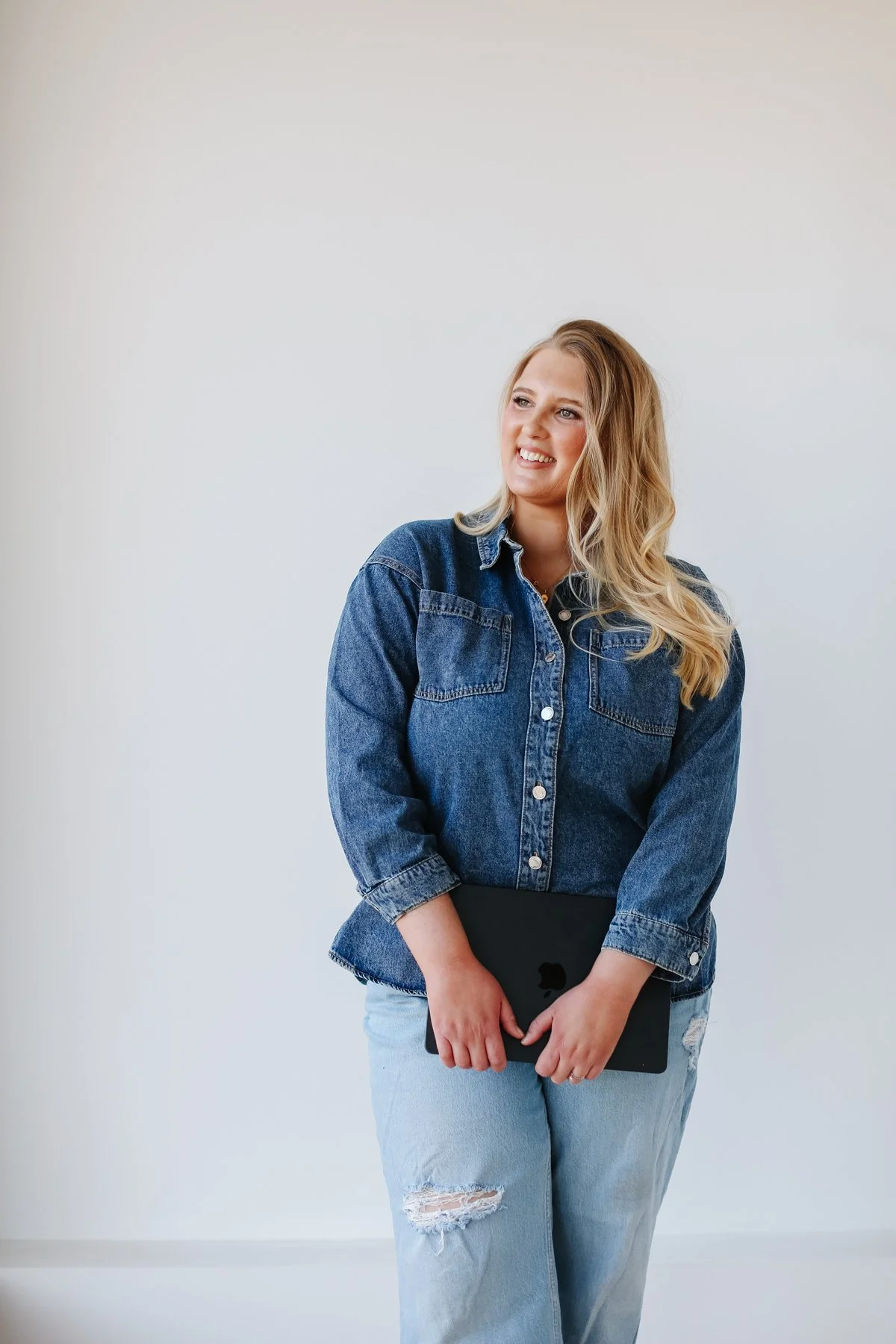 A woman with wavy blonde hair wearing a denim shirt and ripped jeans holds a black tablet while standing against a plain white wall.