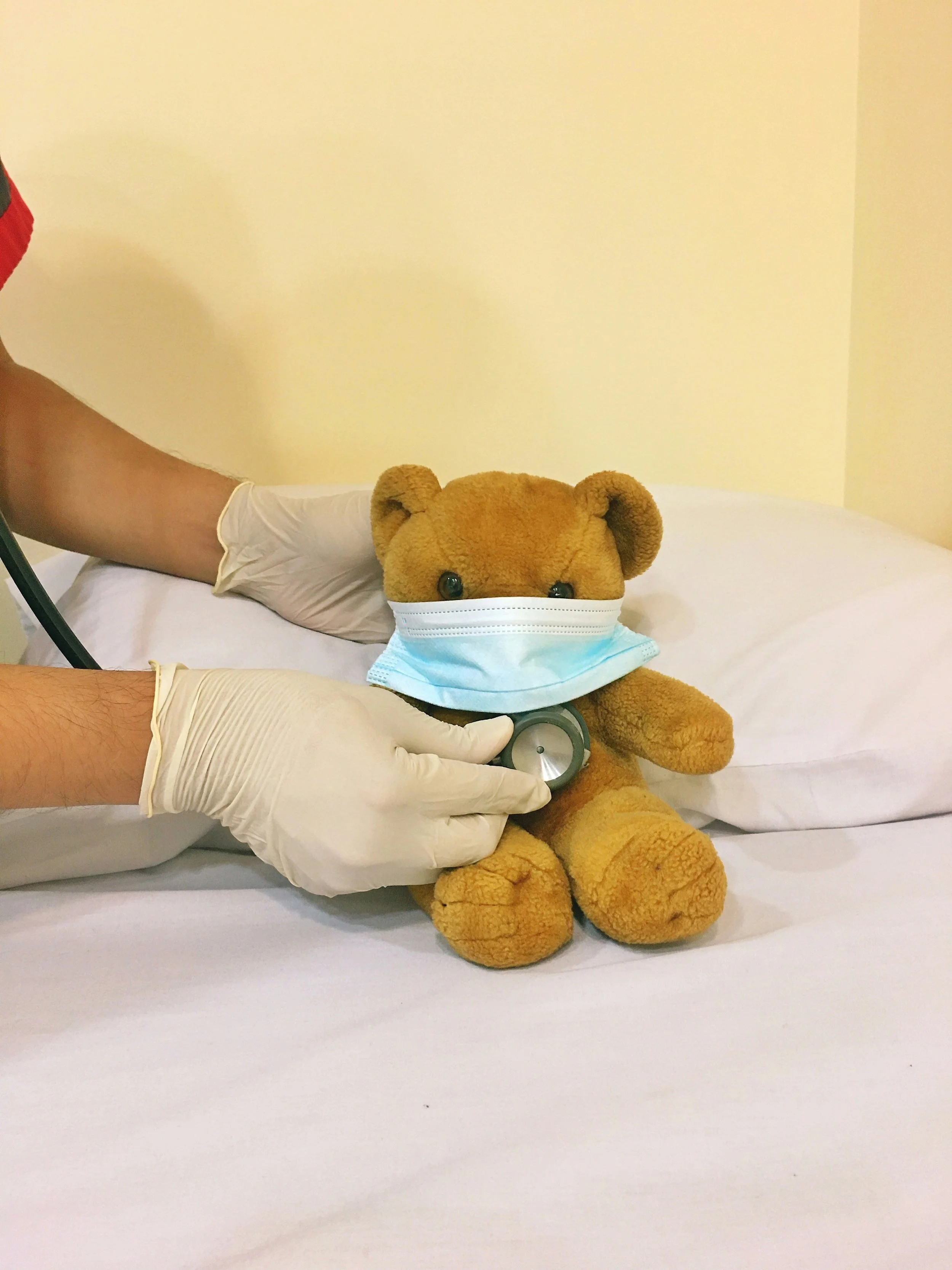 A teddy bear wearing a face mask sits on a hospital bed. A person wearing gloves holds a stethoscope on the bear.