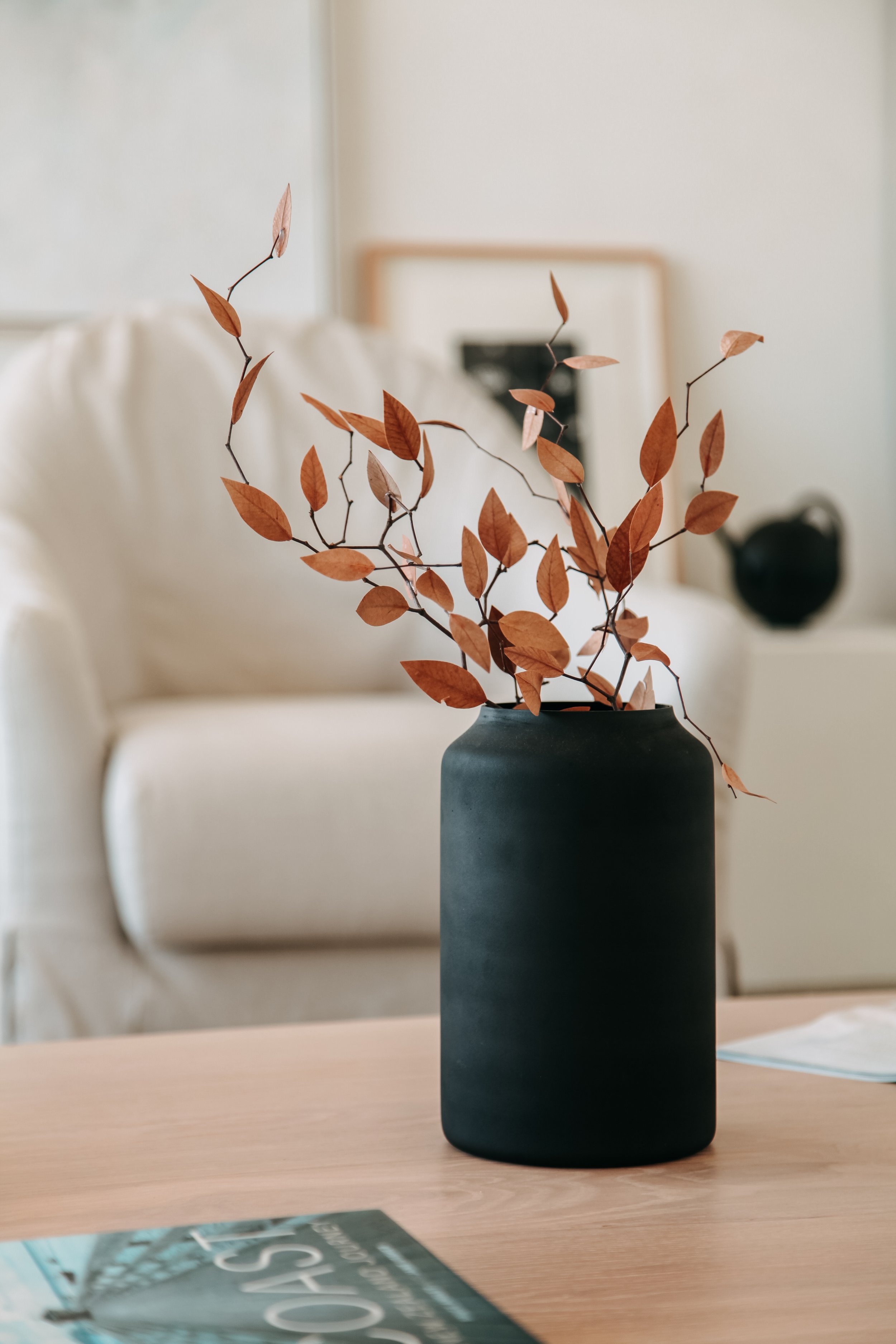 A black ceramic vase with brown leaves on thin, winding branches, placed on a wooden table in a cozy room.