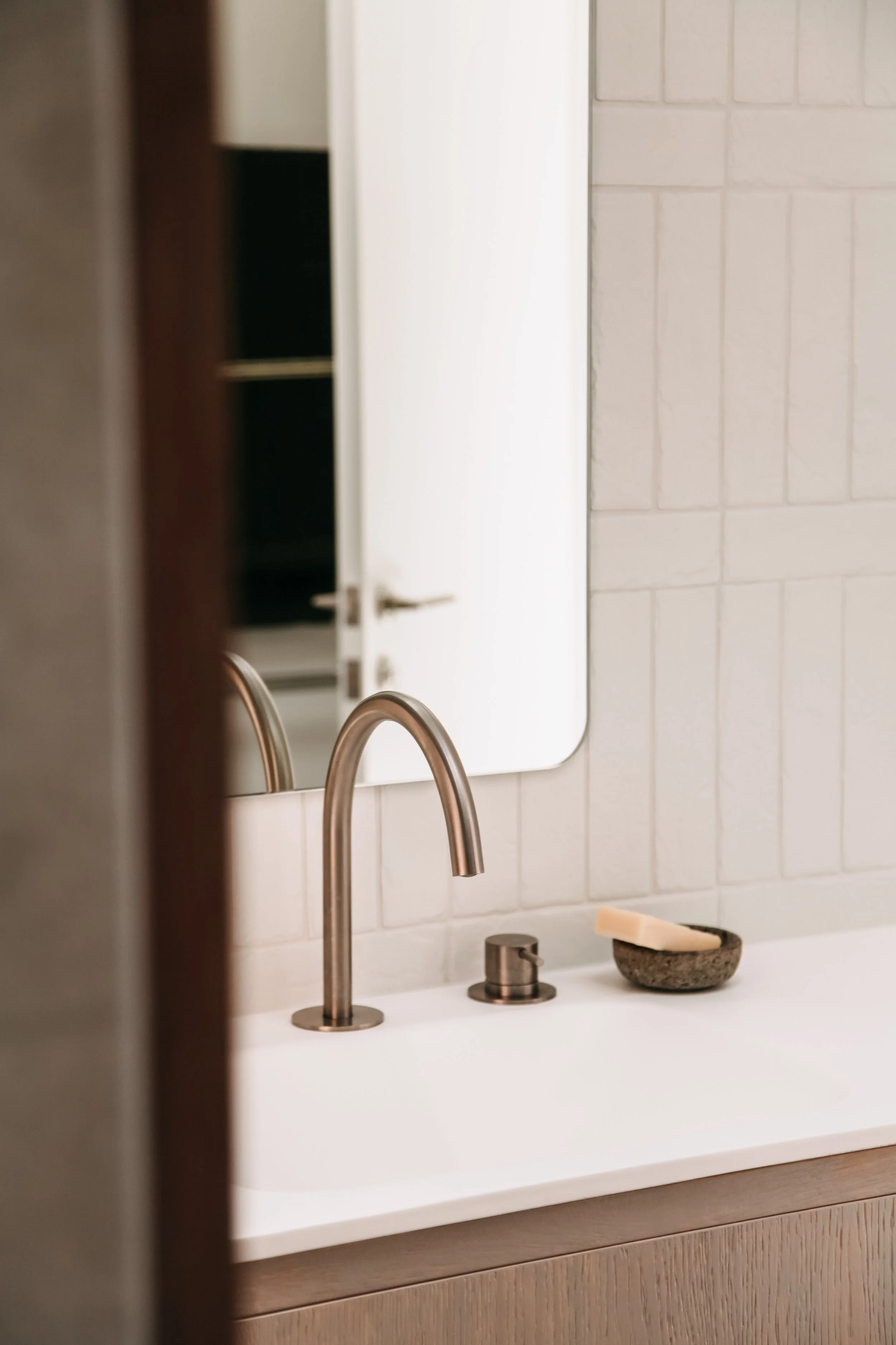 Bathroom sink with a modern faucet, soap in a bowl, a mirror reflecting the door, and tiled wall.