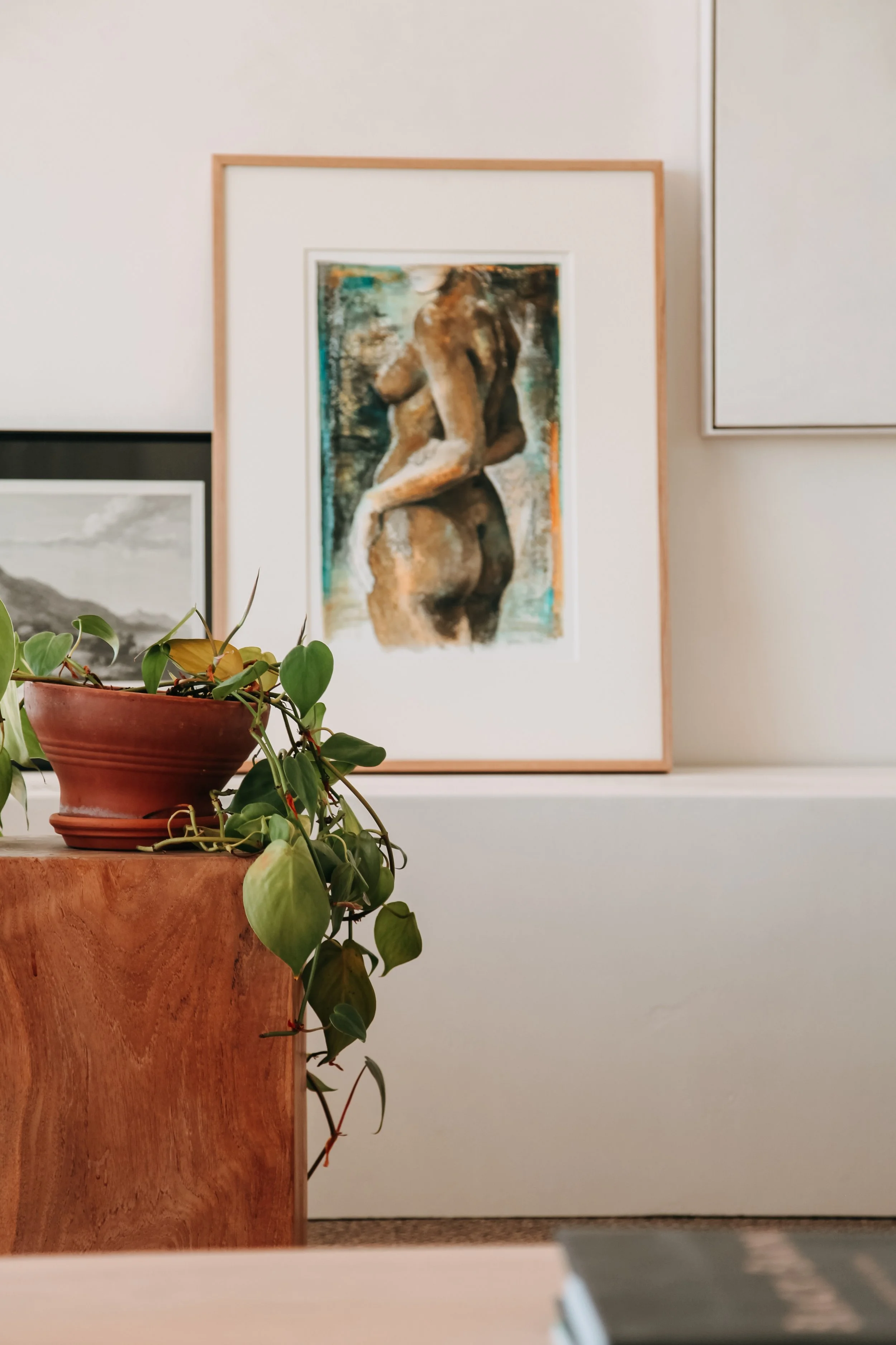 Close-up of a potted plant with trailing green leaves on a wooden surface in front of artwork featuring a painting of a nude woman with back turned, displayed on a gallery wall.