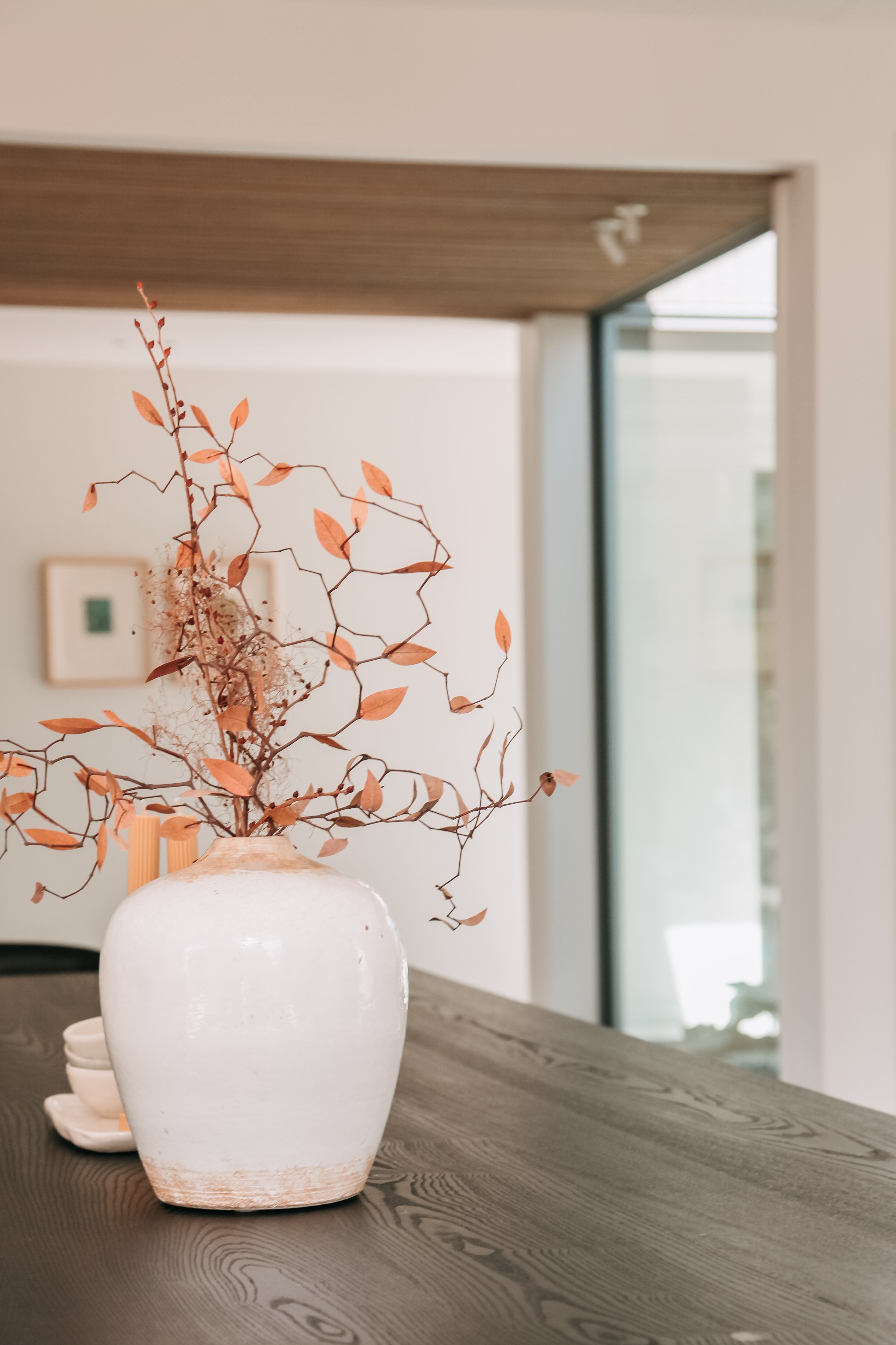 A white ceramic vase with dried peach-colored leaves and twigs, placed on a dark wooden table in a modern interior.