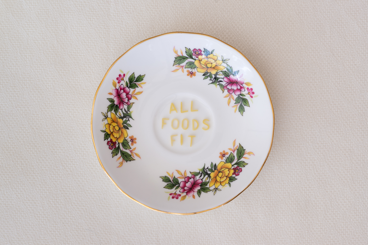 The words All Foods Fit are spelled out in edible pasta letters on a china saucer plate with a dainty pink and yellow floral design. The plate rests on a cream or off-white tablecloth with a subtle woven pattern.