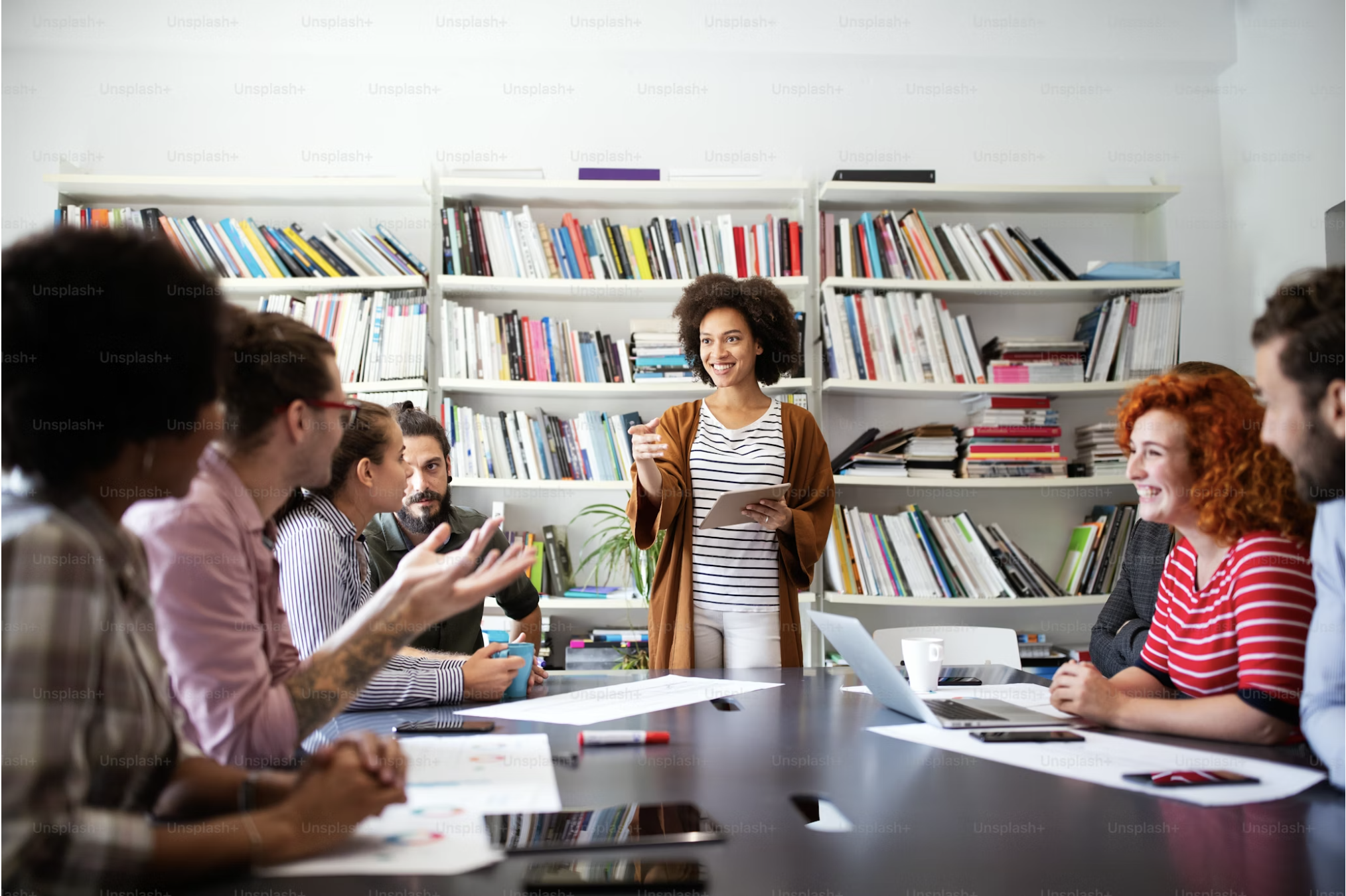 A diverse group of people attending a meeting in a room with bookshelves, with a woman standing and talking to the group.