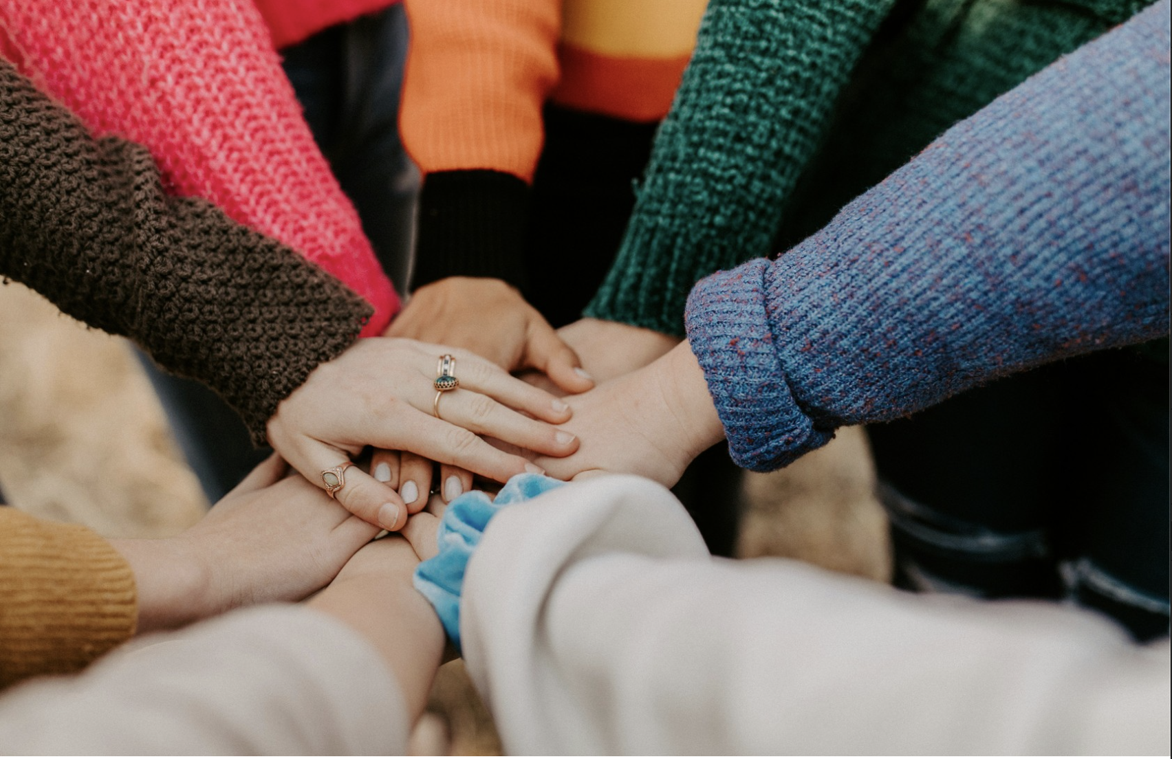A group of people holding hands in a circle, showcasing unity and support. The hands display various rings and are dressed in colorful sweaters.