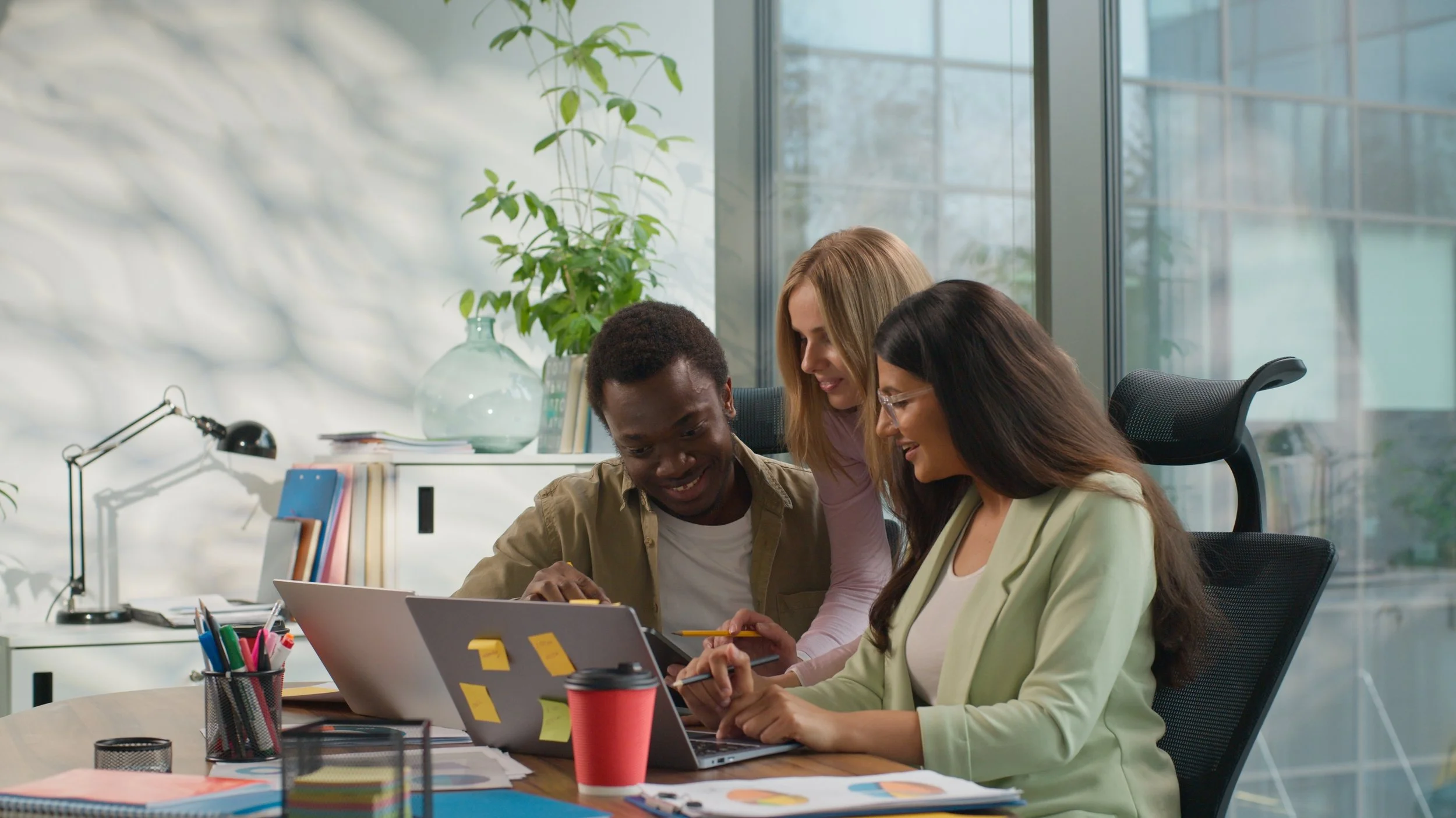 Four diverse colleagues collaborating around a laptop and tablet at a modern office desk, with colorful papers, pens, and coffee cup, in front of large windows with natural light.