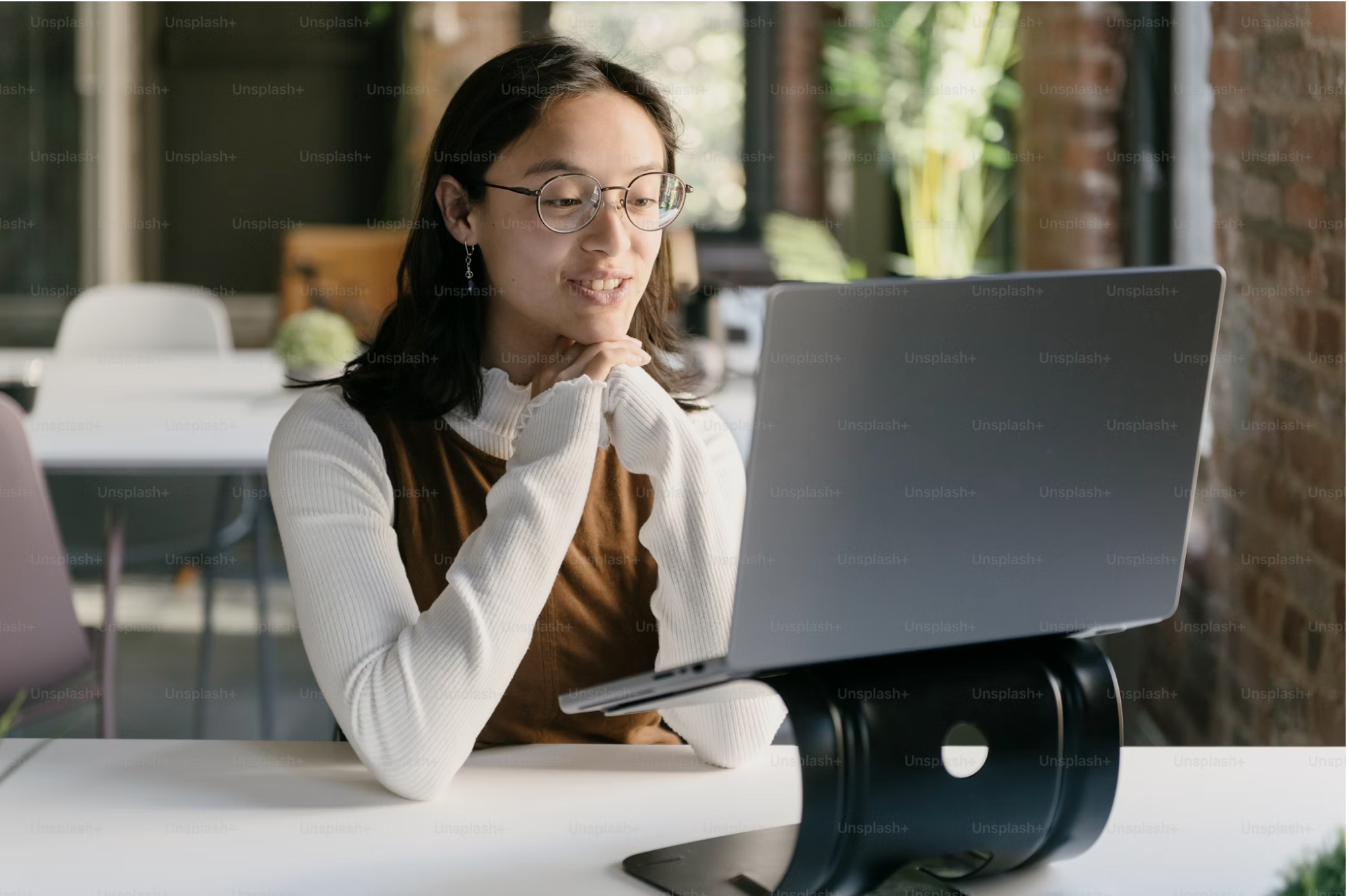 A woman with glasses and earrings sitting at a table, looking at a laptop, in a modern, well-lit room with brick walls and large windows.