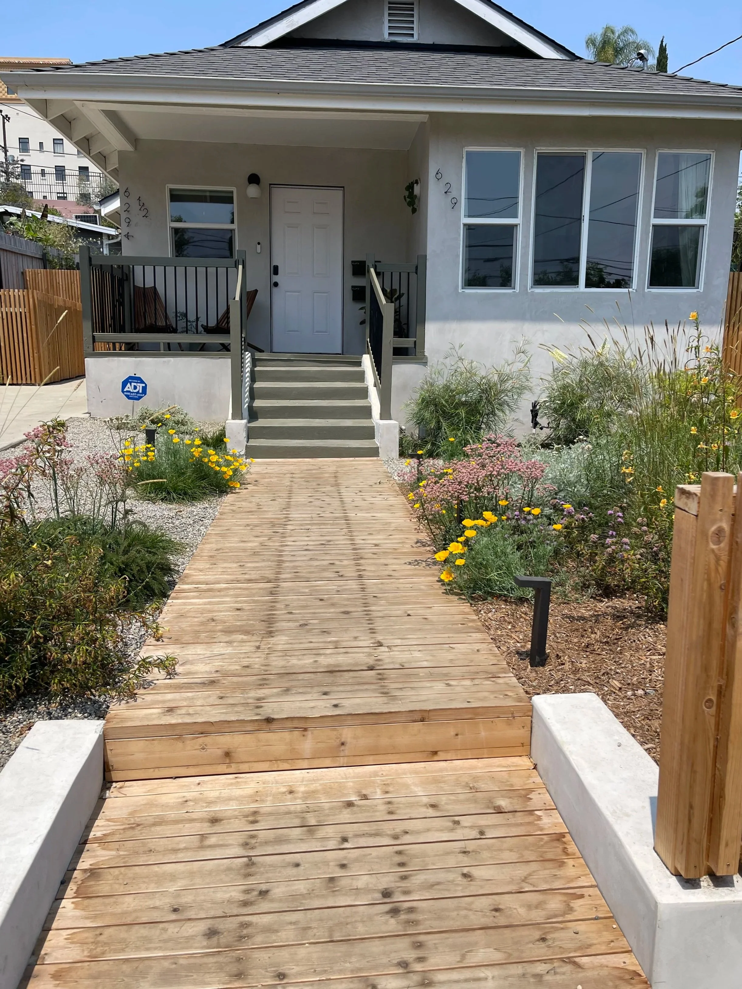 Wood boardwalk ramp leading to a house's front steps and door, flanked by flower beds