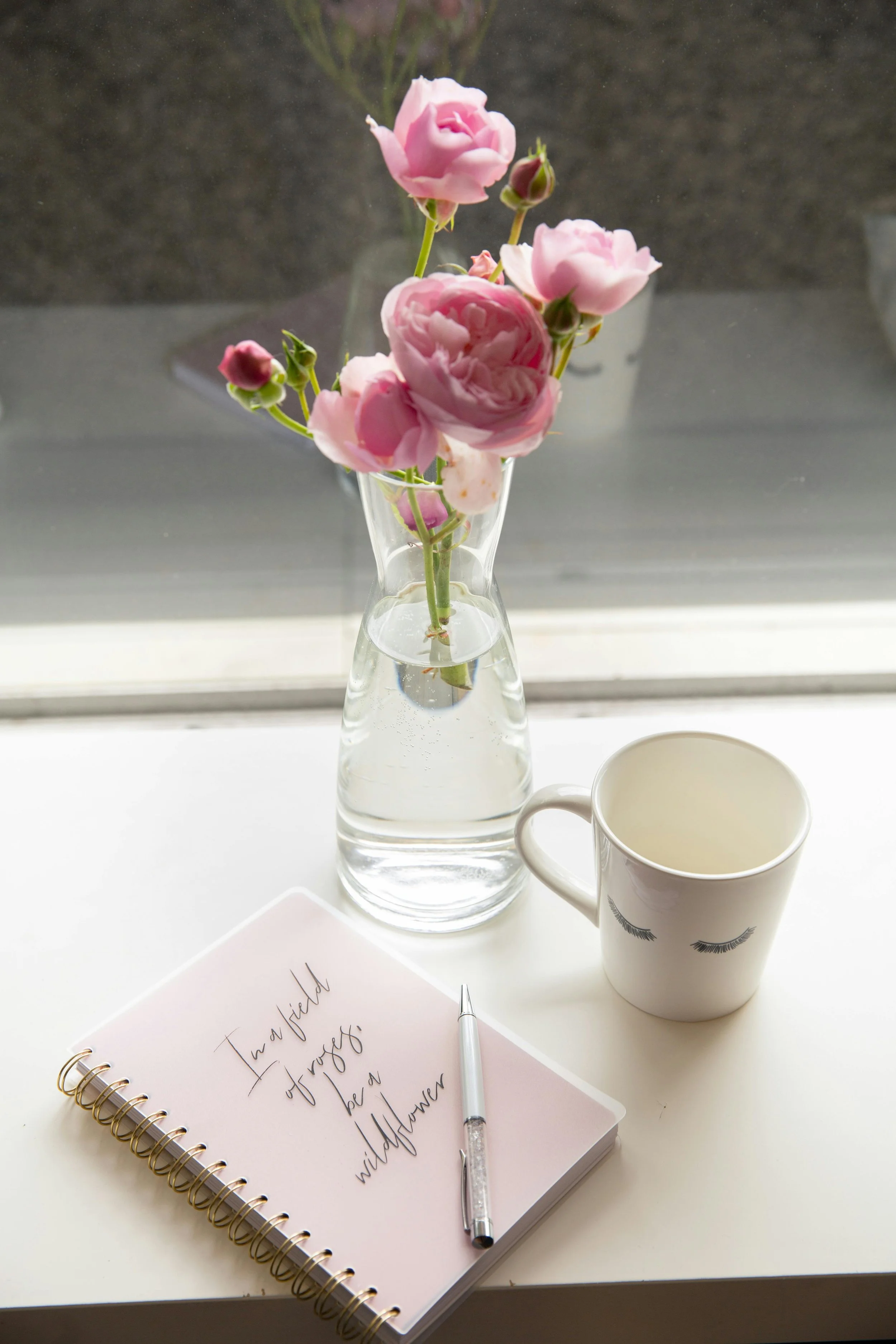 A pink flower arrangement in a glass vase, a white mug with eyelash designs, a pink notebook with handwritten text, and a white pen on a white surface by a window.